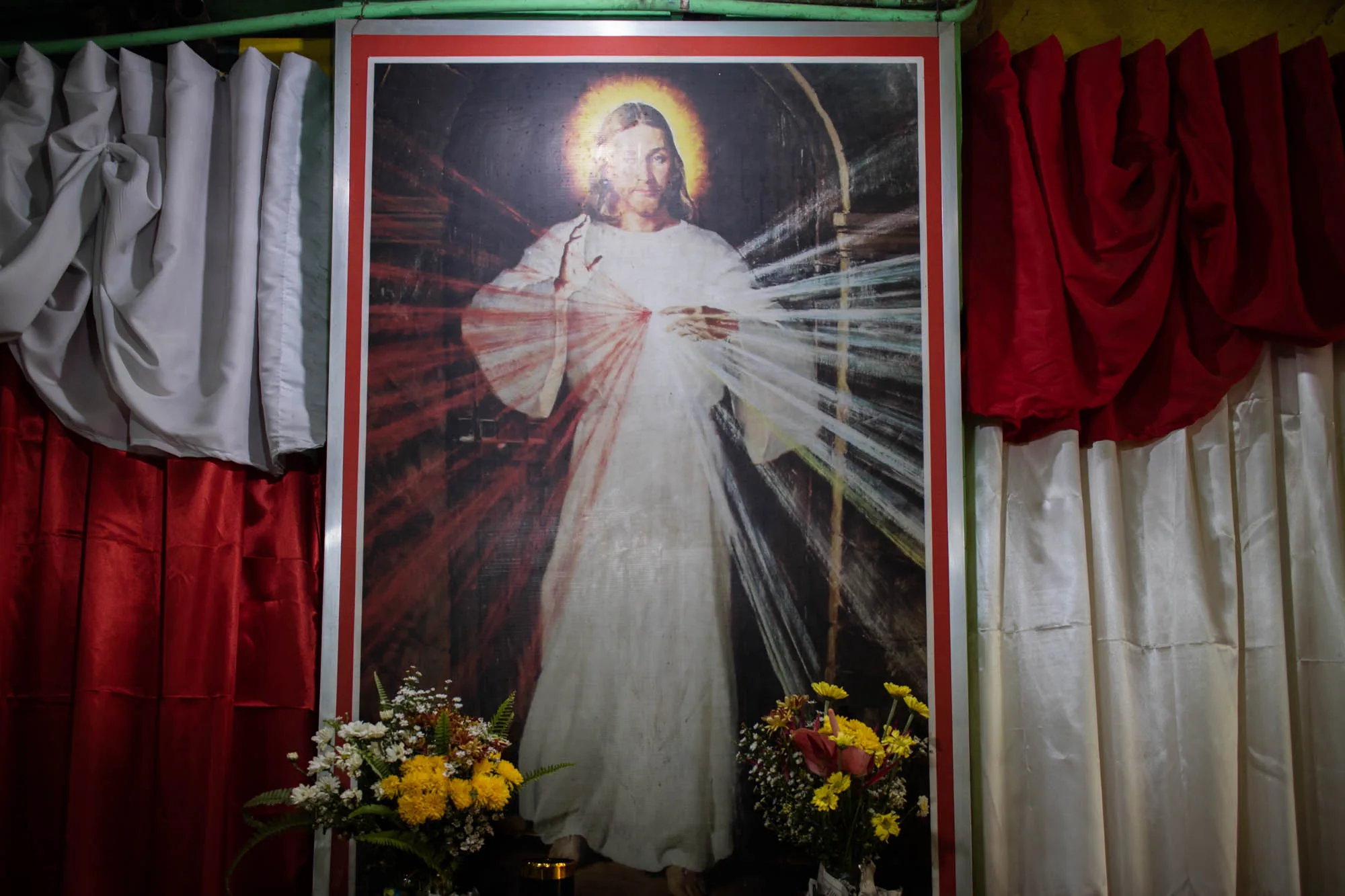 写真には、キリストの絵が飾られています。キリストは白いローブを着て、赤い光線と白い光線が両手から放射されています。頭上には光輪があります。絵の周りには赤と白のカーテンがかけられており、絵の前には黄色や白の花で飾られた花束が2つ置かれています。全体的に、宗教的な雰囲気を感じさせる写真です。場所はフィリピンのセブであることから、信仰心の厚い地域であることが想像できます。