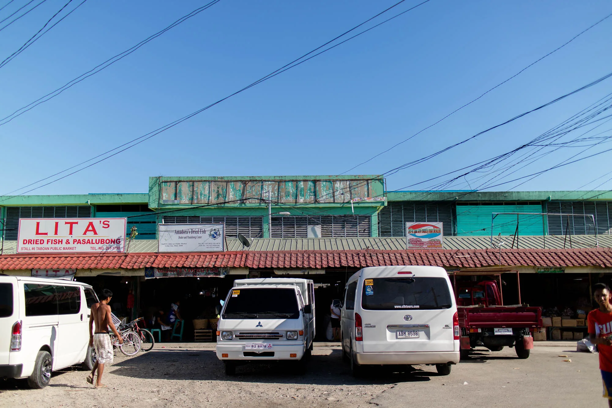 写真はフィリピンのセブで撮影された市場の様子を捉えています。手前には数台の白いバンと赤いトラックが駐車しており、その向こうには市場の建物が見えます。建物は緑色の壁で、屋根は赤茶色の瓦葺きです。建物の看板には「LITA'S DRIED FISH & PASALUBONG」と書かれており、乾燥魚を売る店であることがわかります。他にも「Amadora's Dried Fish」という看板があり、このエリアが乾燥魚の販売で知られていることを示唆しています。  空は明るい青色で、電線が複雑に張り巡らされています。左側には上半身裸の男性が歩いており、地元の生活感が伝わってきます。市場の建物の中には、商品が陳列されている様子が見え、人々が買い物や商売をしている様子もうかがえます。全体的に活気があり、ローカルな雰囲気が漂う写真です。