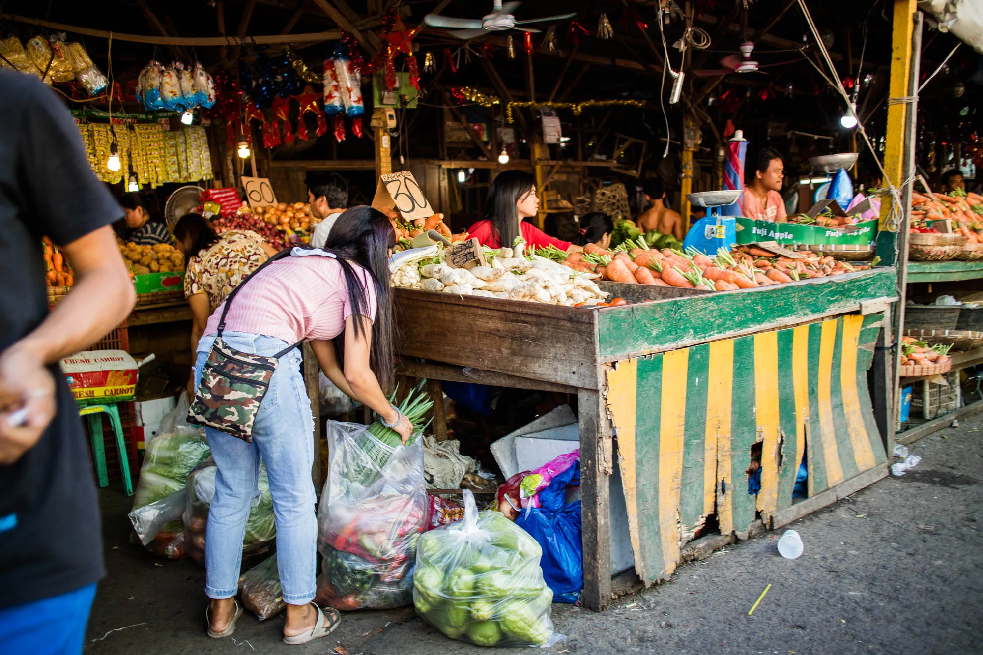 写真はフィリピンのセブで撮影されたもので、活気ある市場の様子が写っています。手前には、ピンク色のトップスとジーンズを身に着けた女性が、野菜の束を手に取ろうと身をかがめています。彼女の足元には、たくさんの野菜が詰まった透明なビニール袋が置かれています。  後ろには、ニンジンやサトイモなどの新鮮な野菜が山積みにされた露店が並んでいます。露店の屋根には電球や装飾品が吊るされ、活気に満ちた雰囲気を醸し出しています。奥には店員や他の買い物客の姿も見え、市場の賑わいが感じられます。露店の前には、商品の値段が書かれた看板が立てられています。全体的に、市場の活気と人々の生活が感じられる一枚です。