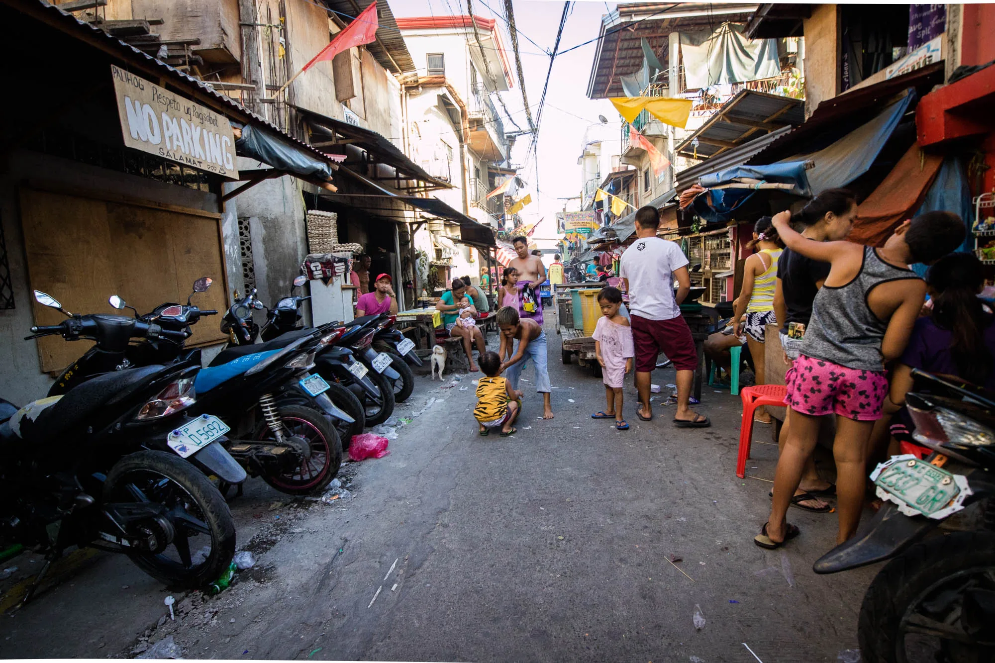この写真はフィリピンのセブで撮影されたもので、路地の生活風景を捉えています。路地は比較的狭く、両側には建物が密集しています。建物の多くは簡素な造りで、屋根はトタンで覆われている箇所もあります。電線が縦横に走り、生活感を感じさせます。  路地には複数の人々がおり、子供たちが遊んでいる姿も見られます。大人は談笑したり、子供の世話をしたりと、リラックスした様子です。路地の脇にはバイクが数台停められており、人々の移動手段として利用されていることが伺えます。  写真全体からは、賑やかで活気のある、セブの庶民の生活が感じられます。