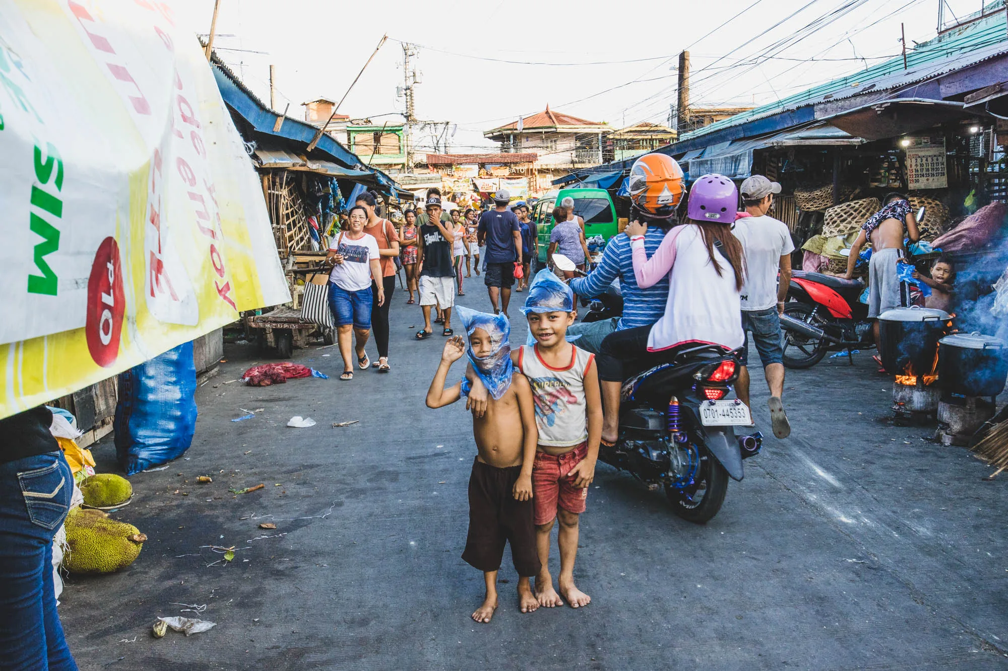 写真はフィリピンのセブで撮影されたもので、活気あふれる市場の様子を捉えています。画面の中央には、ビニール袋を頭にかぶった二人の子供が立っており、一人はカメラに手を振っています。子供たちの後ろには、オレンジと紫のヘルメットを被った人が乗るバイクが停車しています。  周囲には多くの人が行き交っており、日常の喧騒が感じられます。露店が並び、食べ物を調理する煙が立ち上っています。地面はやや汚れ、ジャックフルーツが転がっているのが見えます。建物の構造や人々の服装から、現地の生活感が伝わってきます。全体的に活気があり、生活感あふれる市場の様子を捉えた一枚です。