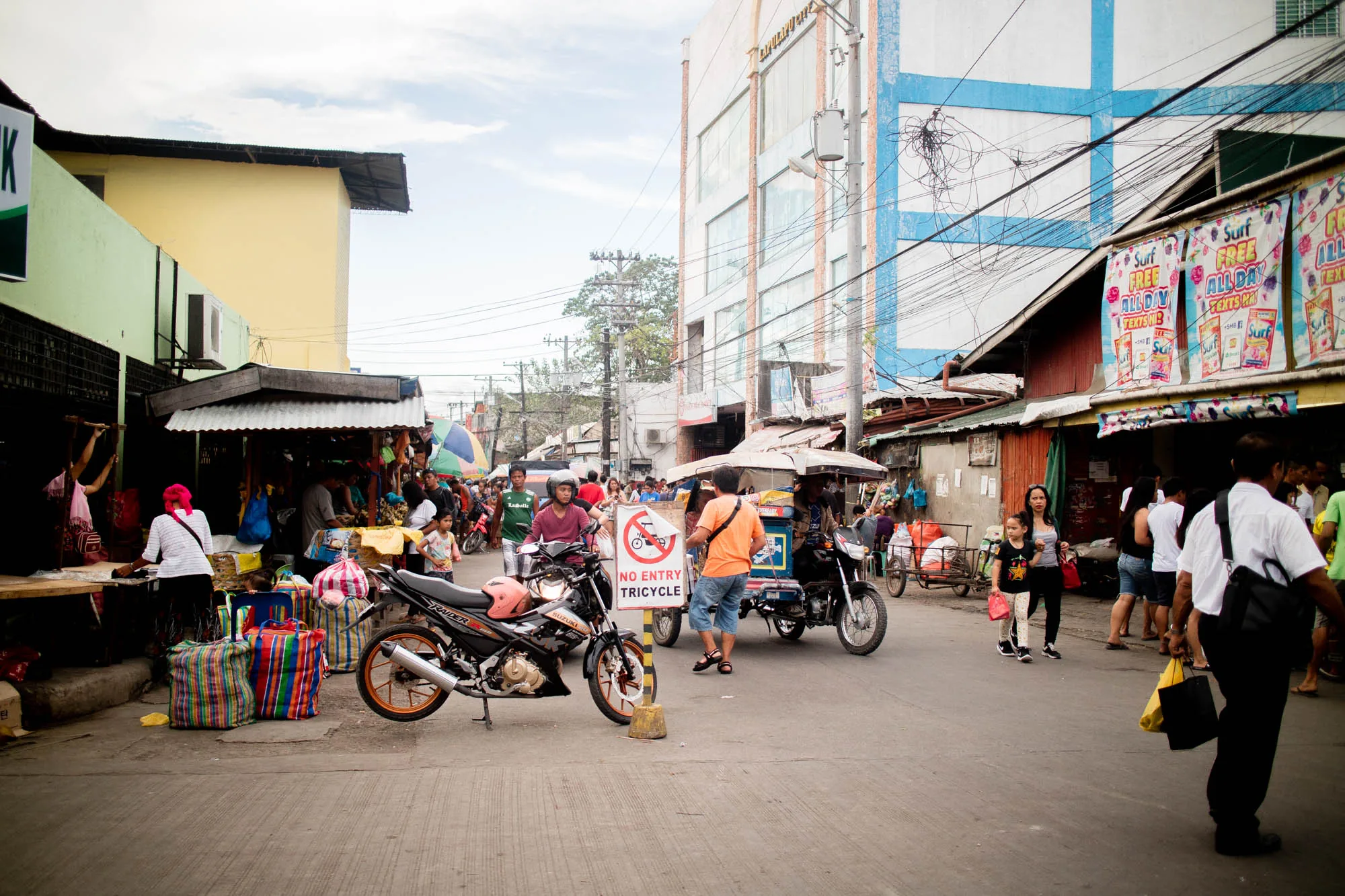 フィリピンのセブで撮影された写真です。路地は市場になっており、多くの人々で賑わっています。手前にはオートバイが停められ、「NO ENTRY TRICYCLE」と書かれた標識があります。奥には、カラフルな傘の下に様々な商品を並べた露店が立ち並び、人々が買い物や行き来をしています。電線が複雑に張り巡らされ、活気あるアジアの市場の雰囲気を醸し出しています。右側には洗剤の広告が貼られた建物が見られます。全体的に少し薄暗いですが、セブの日常の風景を捉えた写真です。