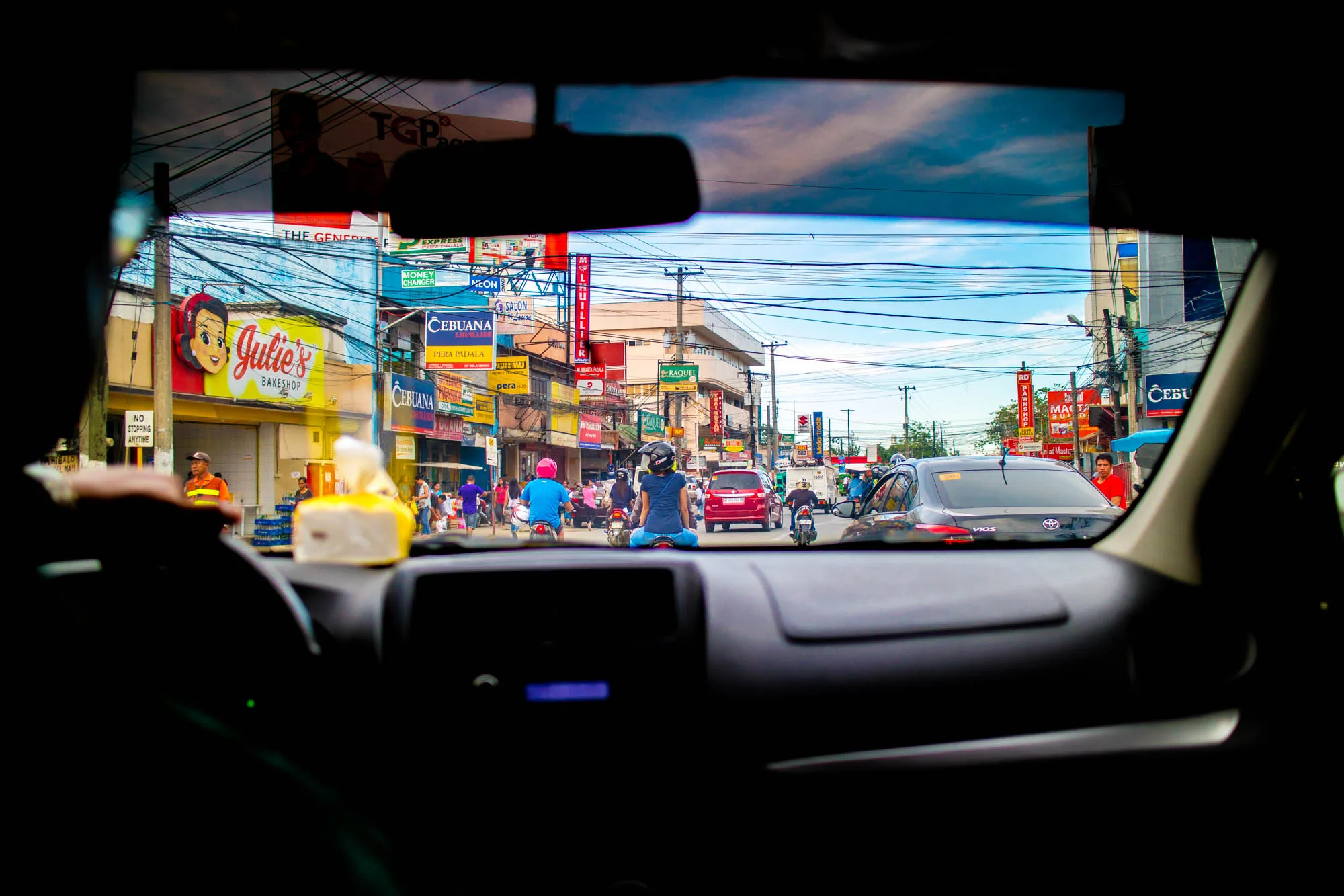 写真は、車のダッシュボード越しに見えるフィリピン、セブの街並みを捉えています。フロントガラスを通して、交通量の多い道路、カラフルな看板が立ち並ぶ建物、そして電線が複雑に張り巡らされた様子が広がっています。左手には「Julie's Bakeshop」というパン屋さんの看板が目立ち、その隣には「NO STOPPING ANYTIME」と書かれた標識があります。道路には、車、バイク、人が行き交い、活気ある雰囲気を醸し出しています。空は青く、一部には雲も見えます。写真全体が、車内からの視点であるため、ダッシュボードが前景となり、フレームのような効果を生み出しています。セブの日常の喧騒とエネルギーが伝わってくる一枚です。