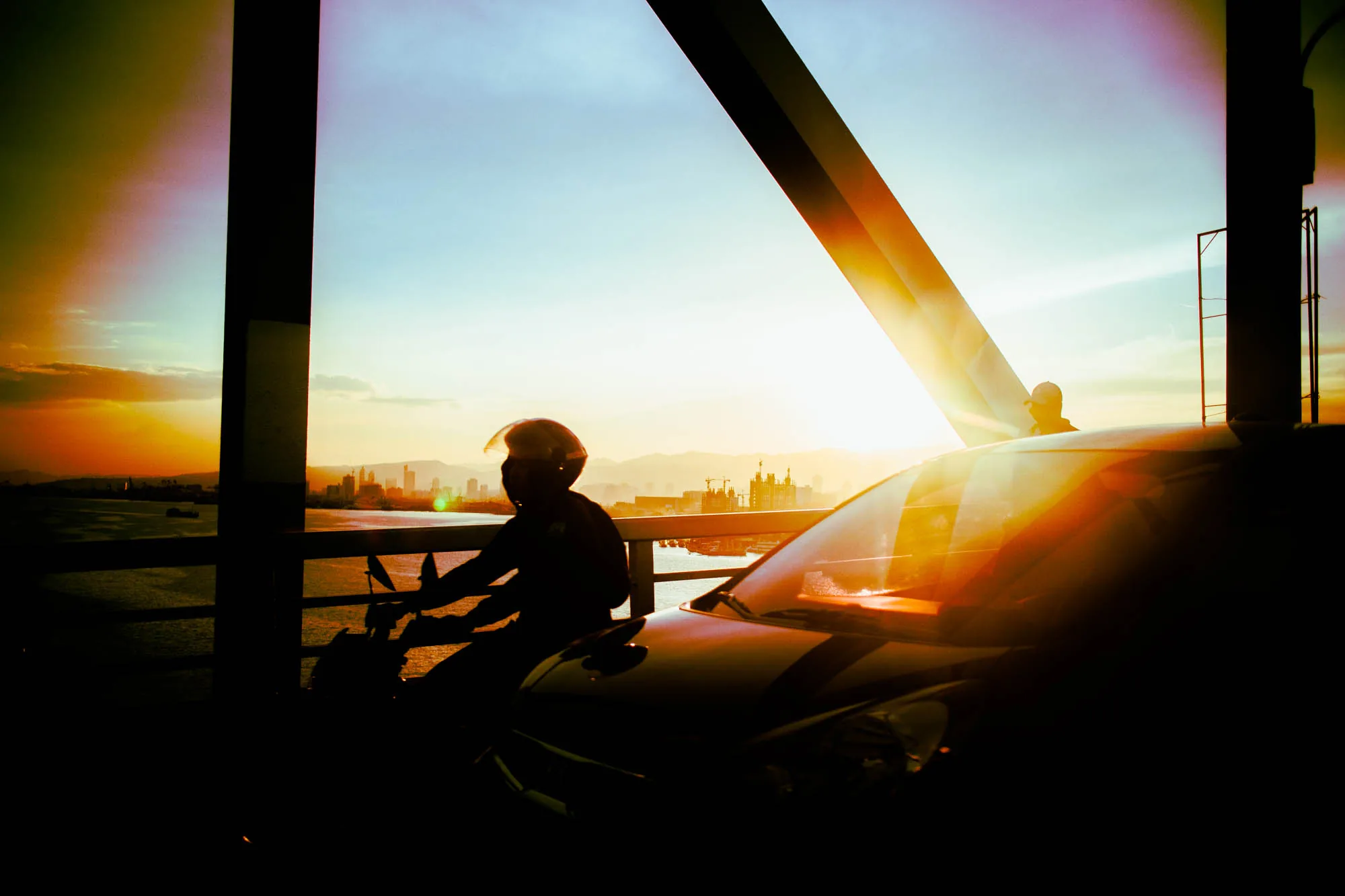 フィリピン、セブで撮影された写真です。夕暮れ時の橋の上で、バイクに乗った人物のシルエットが印象的です。太陽光が画面奥から差し込み、橋の構造物やバイクの車体に反射して、強い光と影のコントラストを生み出しています。背景にはセブの街並みがシルエットで浮かび上がり、海と空が夕焼け色に染まっています。写真全体として、夕暮れ時のセブの美しい風景と、バイクに乗った人物のノスタルジックな雰囲気を捉えています。