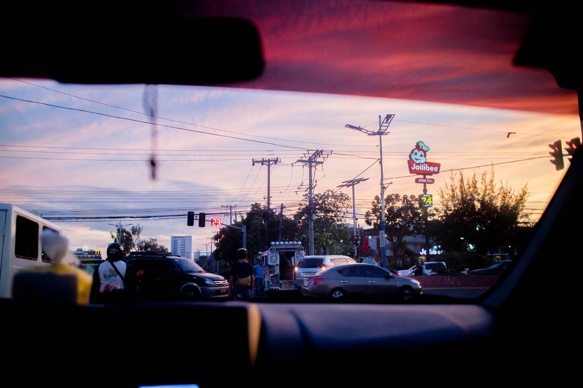 写真はフィリピン、セブの夕暮れ時の交通風景を、車の中から撮影したものです。フロントガラス越しに見える空は、ピンク、オレンジ、紫が混ざり合った美しいグラデーションを描き、夕焼け独特の温かみのある光が街を照らしています。道路にはジープニーや自家用車が行き交い、フィリピンの日常的な交通状況がうかがえます。  右奥には、フィリピンを代表するファストフードチェーン店「Jollibee」の看板が見えます。看板はDrive Thruの文字を示しており、24時間営業していることがわかります。電柱や電線が空を横切り、都市部の風景を象徴しています。手前のダッシュボードは暗く、窓の外の明るさとのコントラストが印象的です。全体的に、夕暮れ時のセブの街の活気と、どこか懐かしい雰囲気を捉えた写真と言えるでしょう。