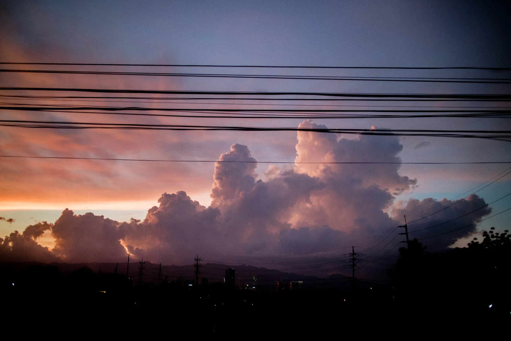 フィリピンのセブで撮影された夕焼け空の写真です。手前には電線が何本も横切っており、シルエットになった街並みと電柱が連なっています。奥には山並みが見え、その上に大きく発達した積乱雲のような雲が浮かんでいます。空はグラデーションになっており、地平線に近い部分はオレンジ色やピンク色、上空は紫色や青色が混ざり合っています。雲も夕焼けの光を受けて、ピンク色や紫色に染まっています。全体的に暗いトーンで、夕暮れ時の静けさや、どこかノスタルジックな雰囲気が感じられます。
