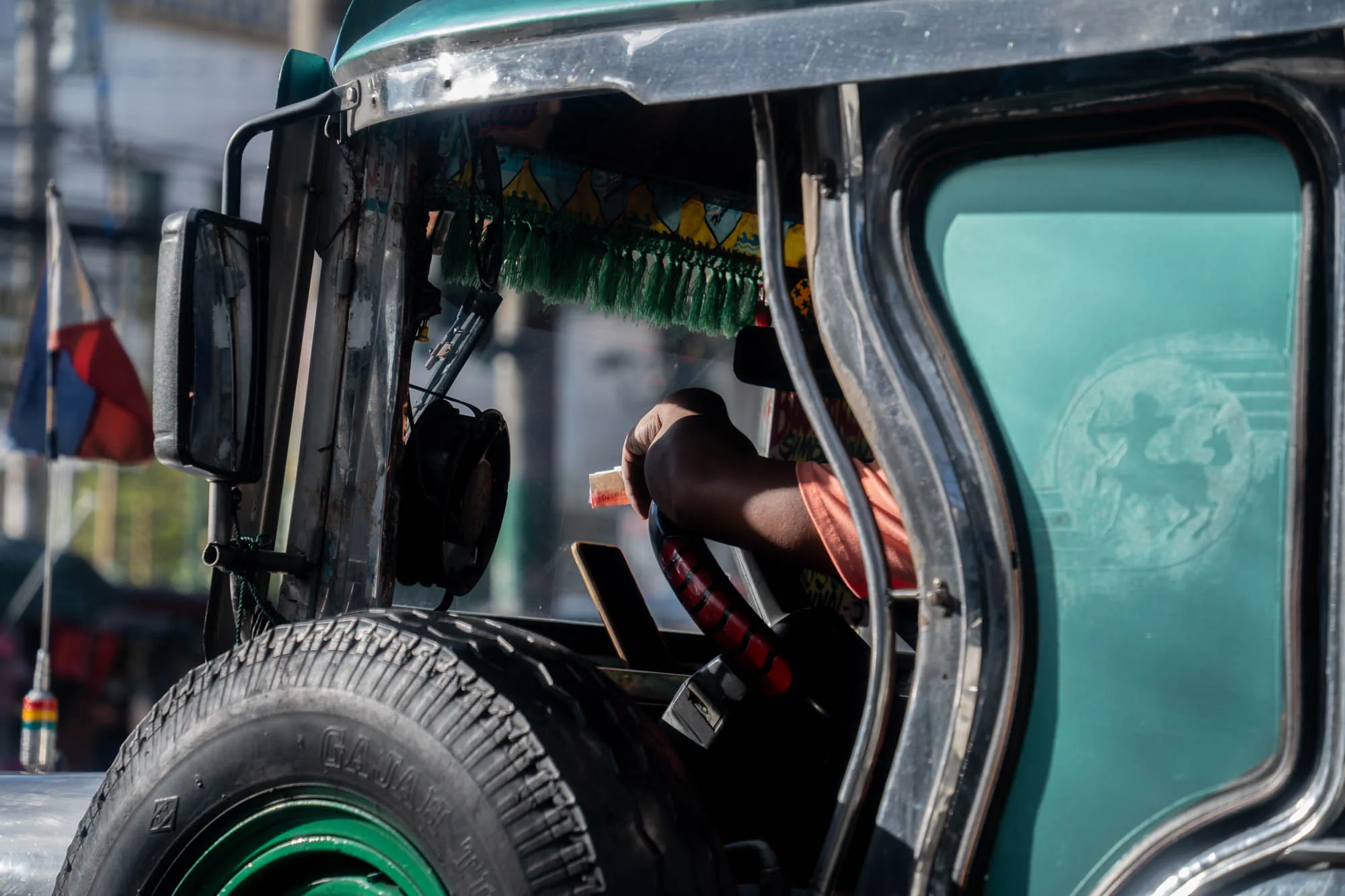 この写真はフィリピンのマニラで撮影された、伝統的な乗り物であるジープニーの運転席付近を捉えたものです。運転手の腕と手、そして赤と黒のステアリングホイールがフレームの中央に位置しています。運転手は何か小さな紙片のようなものを持っています。  ジープニーの外観は、無骨で実用的ながらも装飾的な要素が混在しています。フロントタイヤの一部が見えており、タイヤには「GAJAH」という文字が確認できます。窓ガラスには、人影のような模様がうっすらと浮かび上がっています。  車内には、緑色のフリンジや装飾が施されており、ジープニー特有のカラフルな雰囲気を醸し出しています。バックミラーやフィリピン国旗が背景に見え、マニラの街並みがぼんやりと映り込んでいます。全体的に、ジープニーの独特な個性を際立たせるような、親しみやすい雰囲気の写真です。