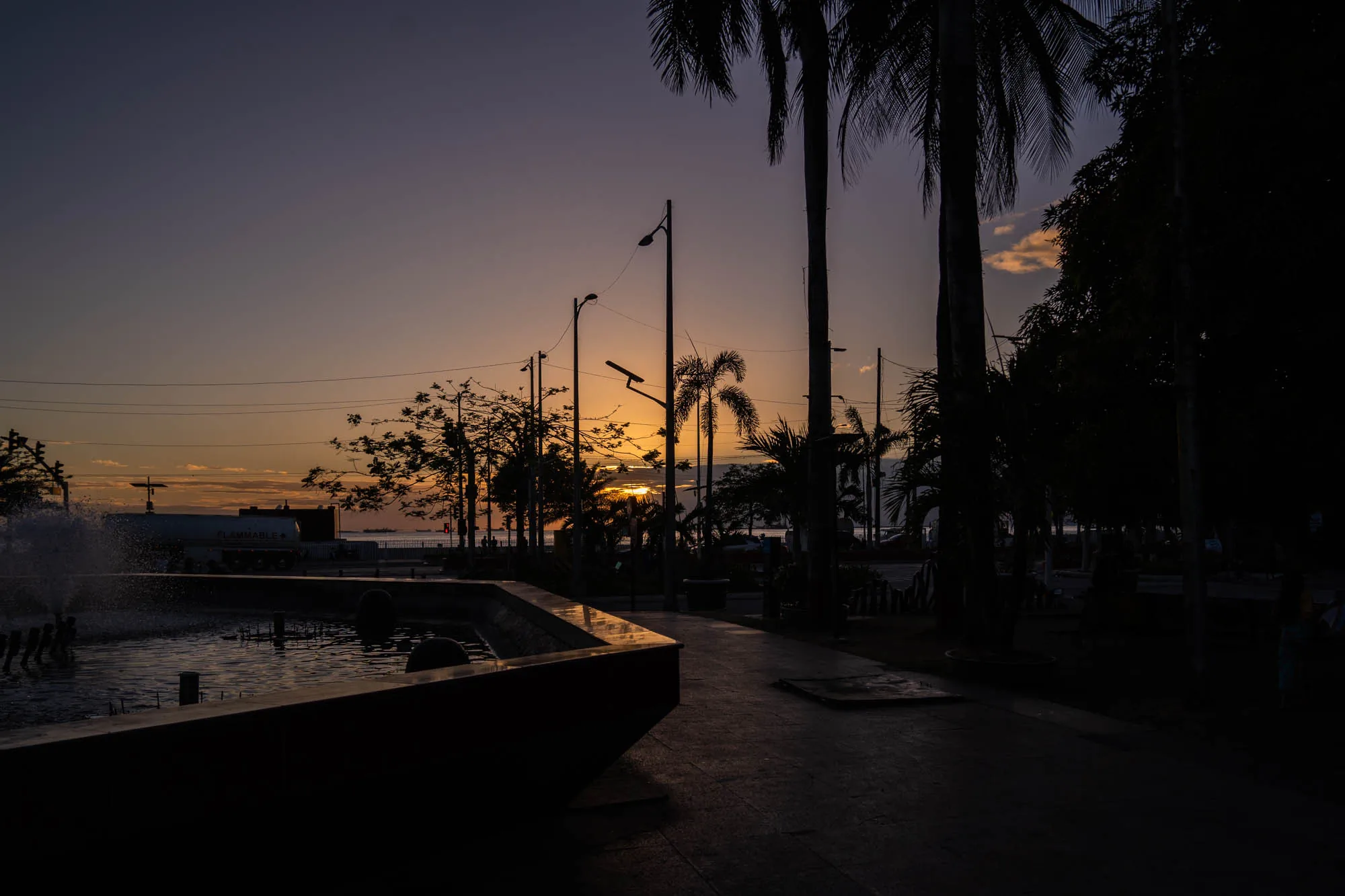 フィリピン、マニラで撮影された夕暮れ時の写真です。手前には噴水があり、水しぶきが上がっています。噴水の向こうには、道路と並木道が続いており、ヤシの木がシルエットになっています。背景には、沈みゆく夕日が空をオレンジ色と紫色に染め上げています。道路にはタンクローリーのような車両も見えます。全体的に暗いトーンで、夕暮れ時の静けさを感じさせる雰囲気です。