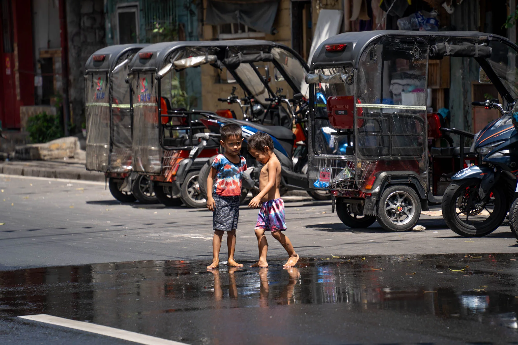 この写真はフィリピンのマニラで撮影されたようです。道路に溜まった水たまりに反射する光が印象的です。水たまりの前に、二人の子供が立っています。一人はタイダイ柄のTシャツとショートパンツを着用し、もう一人は上半身裸で柄物のショートパンツを履いています。裸足で水たまりに足を踏み入れているようです。背景には、フィリピンでよく見られるトライシクルが複数台並んでいます。また、バイクも停まっています。写真全体から、マニラの日常風景と子供たちの無邪気さが伝わってきます。