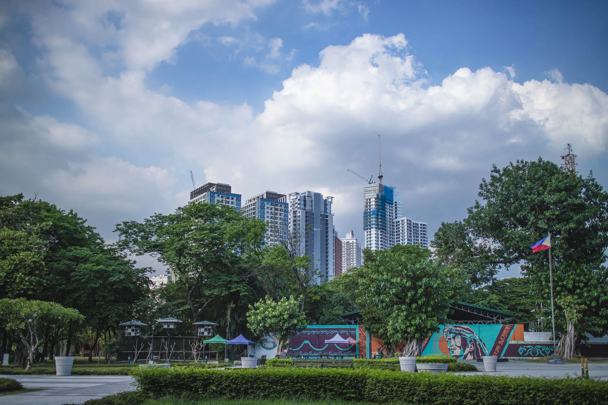 写真はフィリピンのマニラで撮影されたもので、公園の緑と高層ビル群が組み合わさった風景を捉えています。画面奥には、建設中のものも含め、多数の高層ビルが立ち並び、都市開発の進展を示唆しています。空には白い雲が広がり、青空とのコントラストが美しいです。  手前には公園の様子が写っており、緑豊かな木々や整えられた植え込み、遊歩道が見えます。また、フィリピンの国旗が掲げられているのが確認できます。背景には、壁に描かれたカラフルなグラフィティアートが見え、都会的な雰囲気を醸し出しています。公園は整備されており、市民の憩いの場として機能していることが伺えます。全体的に、都市の発展と自然が共存するマニラの様子が伝わってくる写真です。