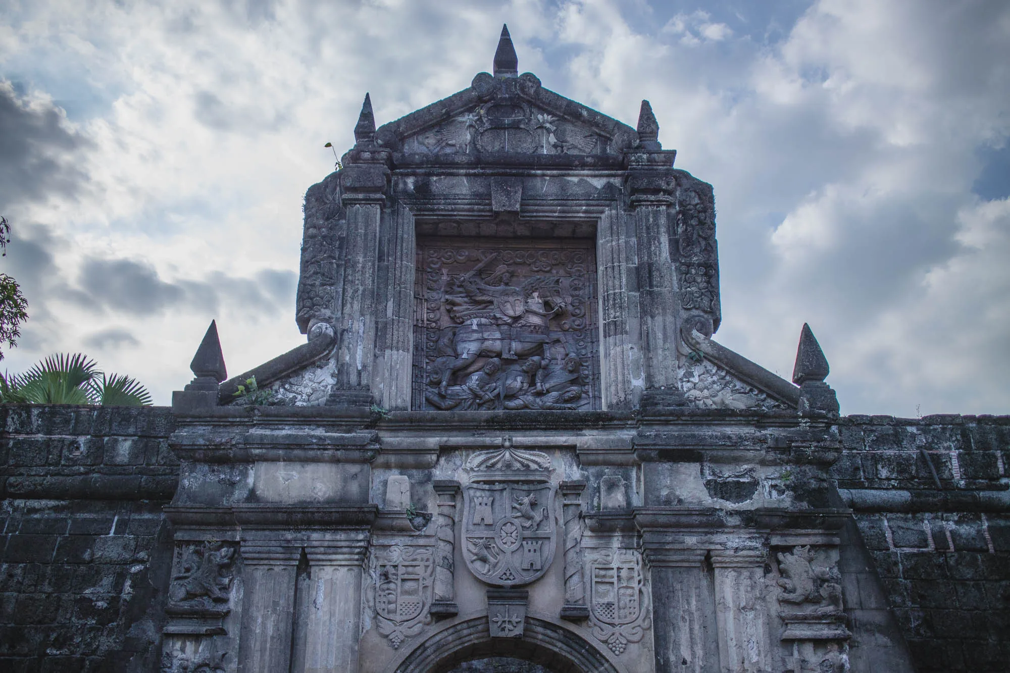 写真はフィリピンのマニラで撮影された、歴史的な建造物の正面を捉えたものです。建物は石造りで、表面は風化し、苔や植物が生えています。上部には騎士が馬に乗って敵を倒している様子を表現した彫刻があり、その下には紋章のようなデザインが施されています。建物の両脇には装飾的な彫刻が施された柱があり、全体的に重厚で歴史を感じさせる雰囲気です。空は曇り空で、建物の陰影を際立たせています。