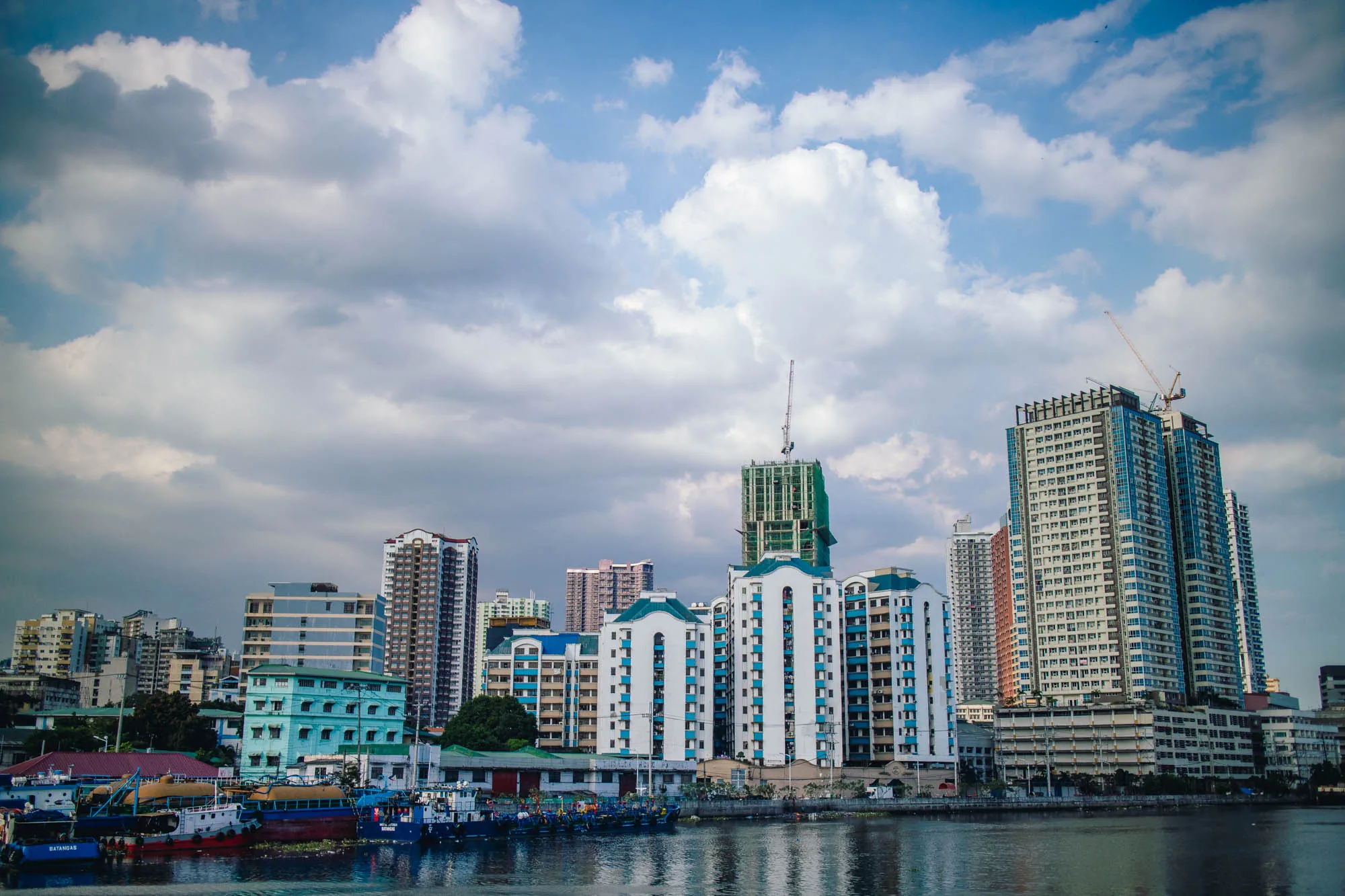 写真はフィリピンのマニラで撮影された都市の景観を捉えています。前景には水面があり、おそらく川または港の一部です。水面には数隻のボートが停泊しており、カラフルな船体が見えます。背景には、様々な高さとデザインの建物が立ち並ぶ市街地が広がっています。特に目を引くのは、白い壁に青いアクセントが施された独特の形状の建物群、建設中の高層ビル、そして複数の高層マンションです。空には厚い雲が広がり、太陽光を遮っているため、全体的にやや落ち着いた印象の写真となっています。空は白と青の濃淡で覆われ、建物群に陰影を与え、都市の活気と穏やかな水辺の風景が組み合わさった様子を描き出しています。