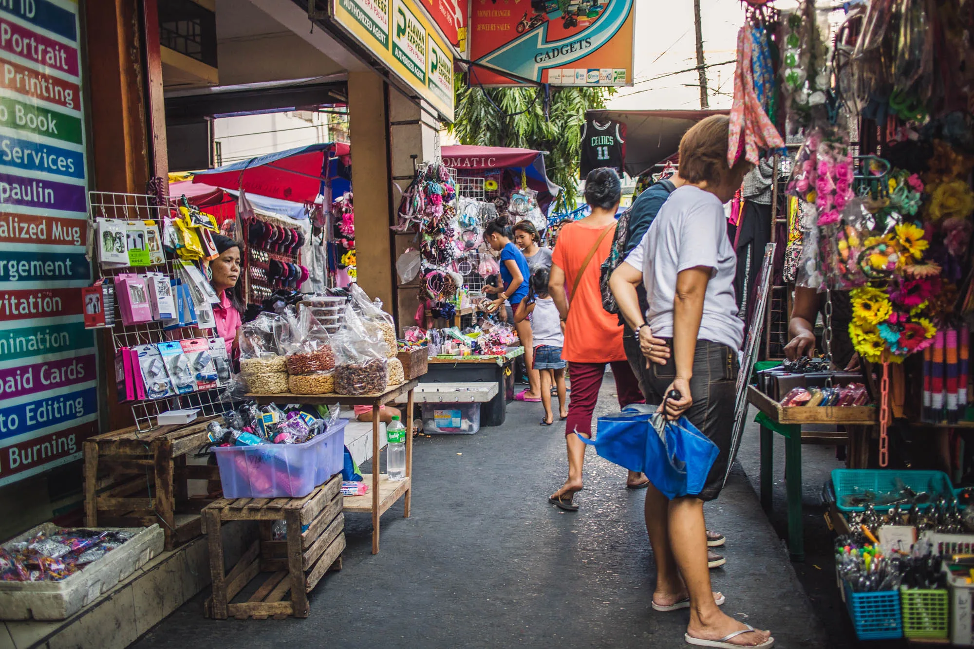 写真はフィリピンのマニラで撮影された市場の様子を捉えています。通り沿いには様々な商品を並べた露店が立ち並び、多くの人々が行き交っています。写真左側の店舗には、「ID Portrait Printing」と書かれた看板が見えます。その他、「alized Mug」「rgement」「itation」「nination」「paid Cards」「to Editing」「Burning」など、様々なサービスの提供を示唆する文字が並んでいます。店先には、イヤホンやアクセサリー、豆菓子などが並べられ、女性店員が接客をしている様子が伺えます。通りの奥には、オレンジ色のシャツを着た女性や子供連れの家族がおり、それぞれ露店の商品を見ています。右側の露店には、花飾りがたくさん飾られており、色鮮やかな雰囲気を醸し出しています。手前の女性は、手に青い傘を持っており、サンダル履きでリラックスした様子です。全体的に活気があり、賑やかな市場の雰囲気が伝わってくる写真です。