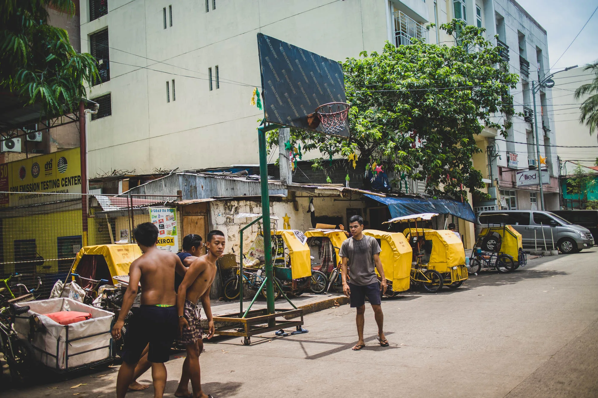 写真はフィリピンのマニラで撮影されたもので、街の日常風景を捉えています。アスファルトの道に、上半身裸の若者たちが数人立っています。そのうちの一人はTシャツと短パン、サンダル姿です。彼らの後ろには、手作りのバスケットボールのゴールがあります。ゴールのバックボードには「MULTIFORM」という文字が繰り返し印刷されています。  背景には、トタン屋根の家々が密集しており、その手前には黄色い幌をつけたトライシクルが数台並んでいます。道の反対側には、Emission Testing Centerの看板がある建物や、他の商業施設の建物が見えます。また、グレーのバンが駐車しています。全体的に、ローカルな雰囲気と生活感が漂う一枚です。画面全体に、日差しによるコントラストが見られます。