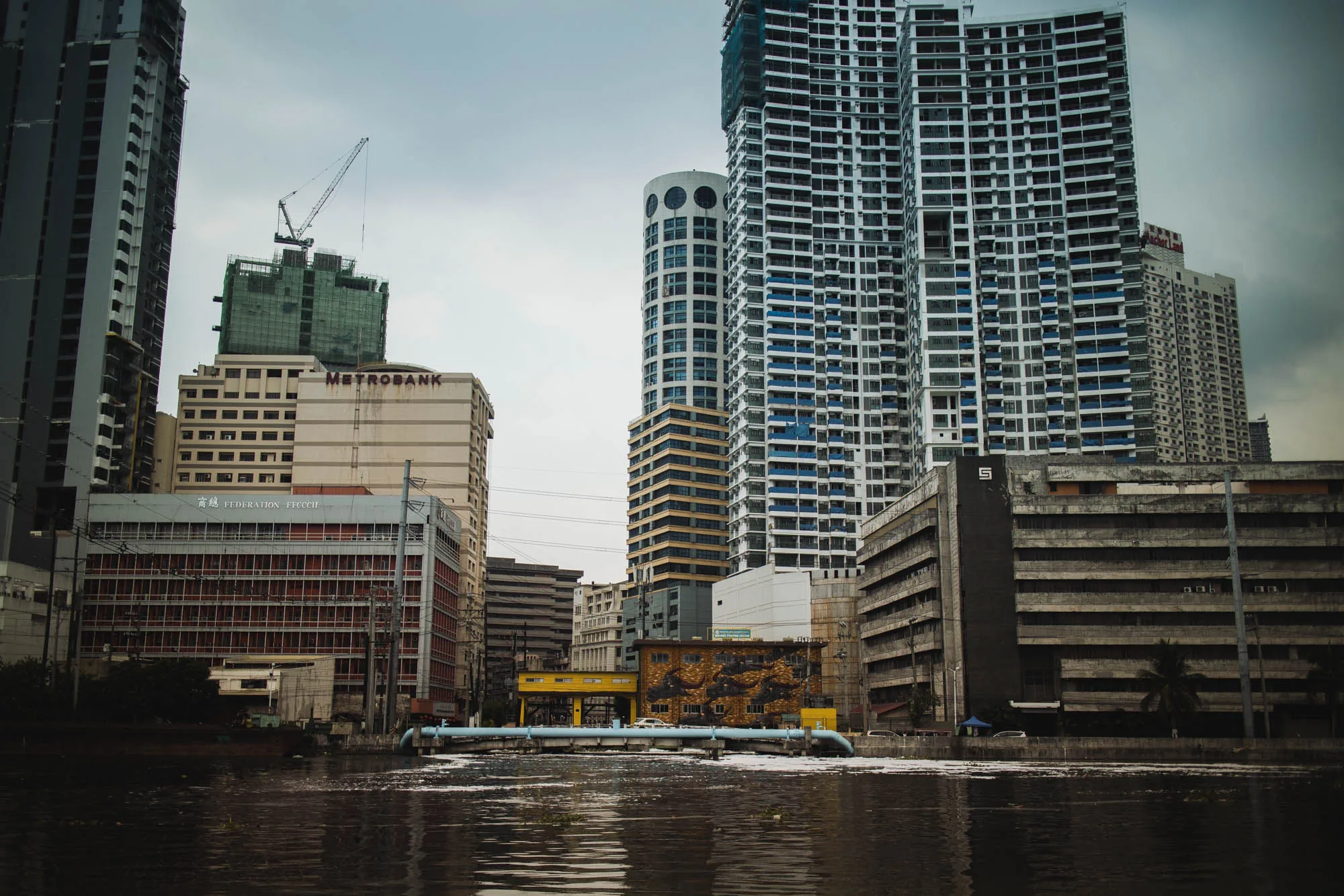 写真は、フィリピンのマニラで撮影された都市の風景を捉えています。前景には、汚れた川が広がり、水面には光が反射しています。川を横切るように、青色のパイプラインが設置されています。  背景には、様々な高さやデザインの建物が密集して立ち並んでいます。高層ビル群は、灰色がかった空の下で、ややくすんだ印象を与えています。その中には、「METROBANK」と書かれた建物や、建設中のクレーンが確認できるビルもあります。また、赤色の窓枠が特徴的な建物や、特徴的な円筒形のデザインを持つビルも目に留まります。  全体的に、写真からは活気のある都市の様子が伝わってきますが、同時に、水質汚染や建物の老朽化といった問題も垣間見えます。都会の喧騒と、それを取り巻く環境の課題が混在した風景と言えるでしょう。