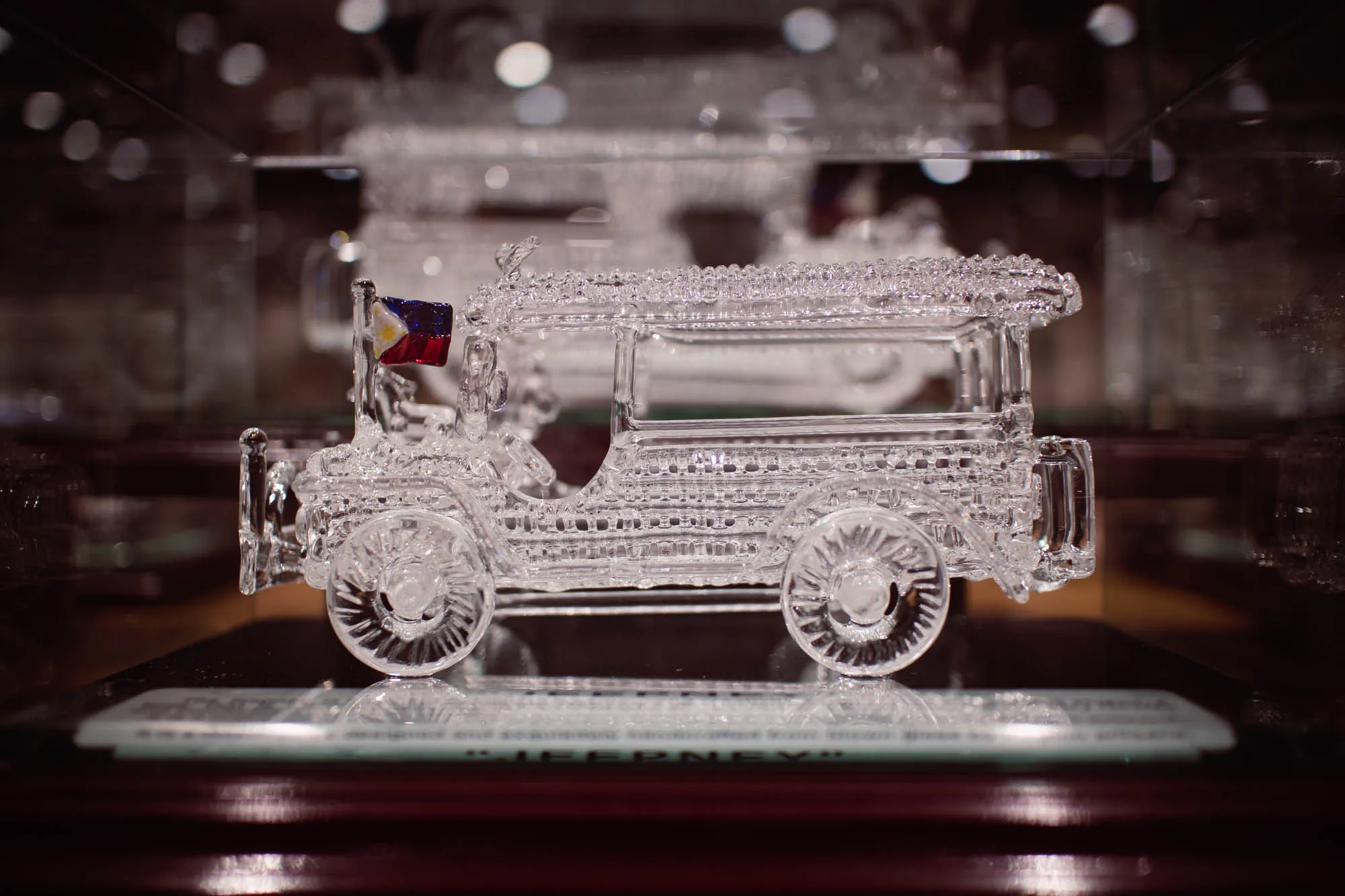 マニラで撮影された写真には、クリスタル製のジープニーの置物が写っています。ジープニーはガラスのケースの中に展示されており、光が反射して輝きを増しています。細部まで丁寧に作られており、車輪、座席、屋根などがクリアに表現されています。フロント部分には小さなフィリピン国旗が取り付けられています。置かれている台座には「JEEPNEY」という文字が見えます。背景はぼやけていますが、暗めの木製のような素材で、高級感のある雰囲気を醸し出しています。全体的に、この写真はフィリピンの文化的なシンボルであるジープニーを、洗練された形で表現した芸術作品のようです。