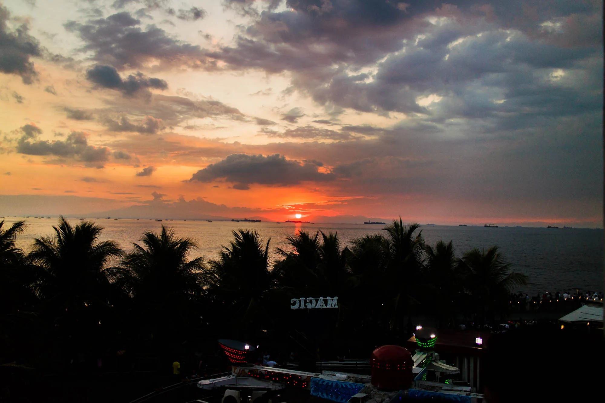 フィリピンのマニラで撮影された夕焼けの風景写真です。画面奥には、太陽が水平線に沈みかけ、空はオレンジ、ピンク、紫といった色で染め上げられています。雲が空全体を覆い、ドラマチックな雰囲気を演出しています。海には複数の船が停泊しているのが見えます。手前にはヤシの木のシルエットが並び、その下には遊園地のような施設が見えます。「MAGIC」と書かれた看板も確認できます。全体的に暗い色調で、夕暮れ時のノスタルジックな雰囲気が漂っています。