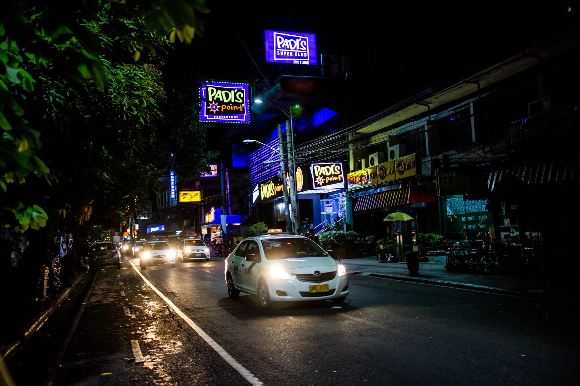 フィリピン、マニラの夜の街並みを捉えた写真です。画面中央には白いタクシーがヘッドライトを光らせながら走行しています。道路は濡れており、タクシーのライトが反射して光沢を放っています。道路の左側には緑の木々が生い茂っており、画面の端まで伸びています。道路の右側には建物が立ち並び、様々なネオンサインが輝いています。"PADI'S Point Restaurant"、"PADI'S SUPER CLUB 2ND FLOOR"といった看板が目を引きます。建物の下にはカフェやレストランらしき店があり、傘の立てかけられた様子も見られます。全体的に暗い夜の雰囲気ですが、ネオンサインや車のライトが活気を与えています。