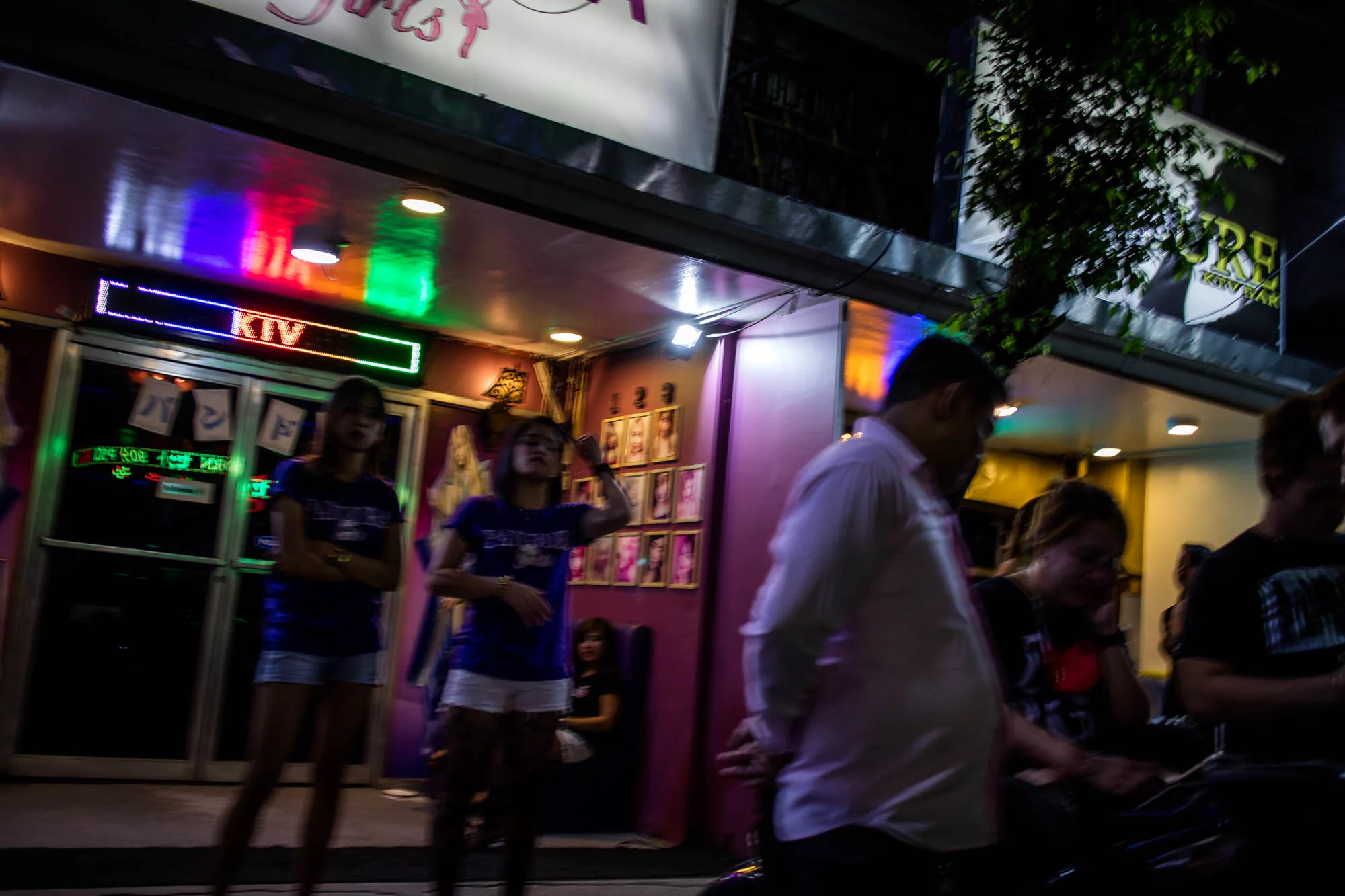 写真はフィリピンのマニラで撮影された夜の風景を捉えています。画面奥には「KTV」と書かれたネオンサインが光る店の入り口があり、店内には複数の女性が確認できます。入り口付近には、短パンとTシャツ姿の女性が立っており、客引きをしている様子が伺えます。店の壁には、女性たちの写真が飾られています。手前には、男性数人が立っており、店に興味を示しているかもしれません。全体的に暗い写真ですが、ネオンサインや街灯がカラフルに輝き、活気のある雰囲気を醸し出しています。猥雑な街の雰囲気が伝わってくる一枚です。