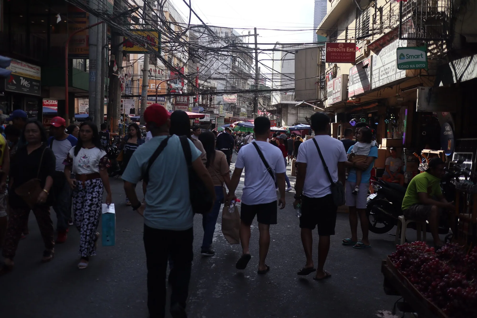この写真はフィリピン、マニラの街中の様子を捉えています。混雑した通りには多くの人々が行き交い、活気にあふれています。電線が複雑に絡み合い、空を覆うように張り巡らされているのが印象的です。建物の壁には看板や広告が所狭しと並び、アジアらしい賑やかさを感じさせます。道の両脇には様々な商店が軒を連ねているのが見て取れます。露店では果物が売られており、人々の日常が垣間見えます。全体的に暗めの写真ですが、人々の喧騒とエネルギーが伝わってくる一枚です。