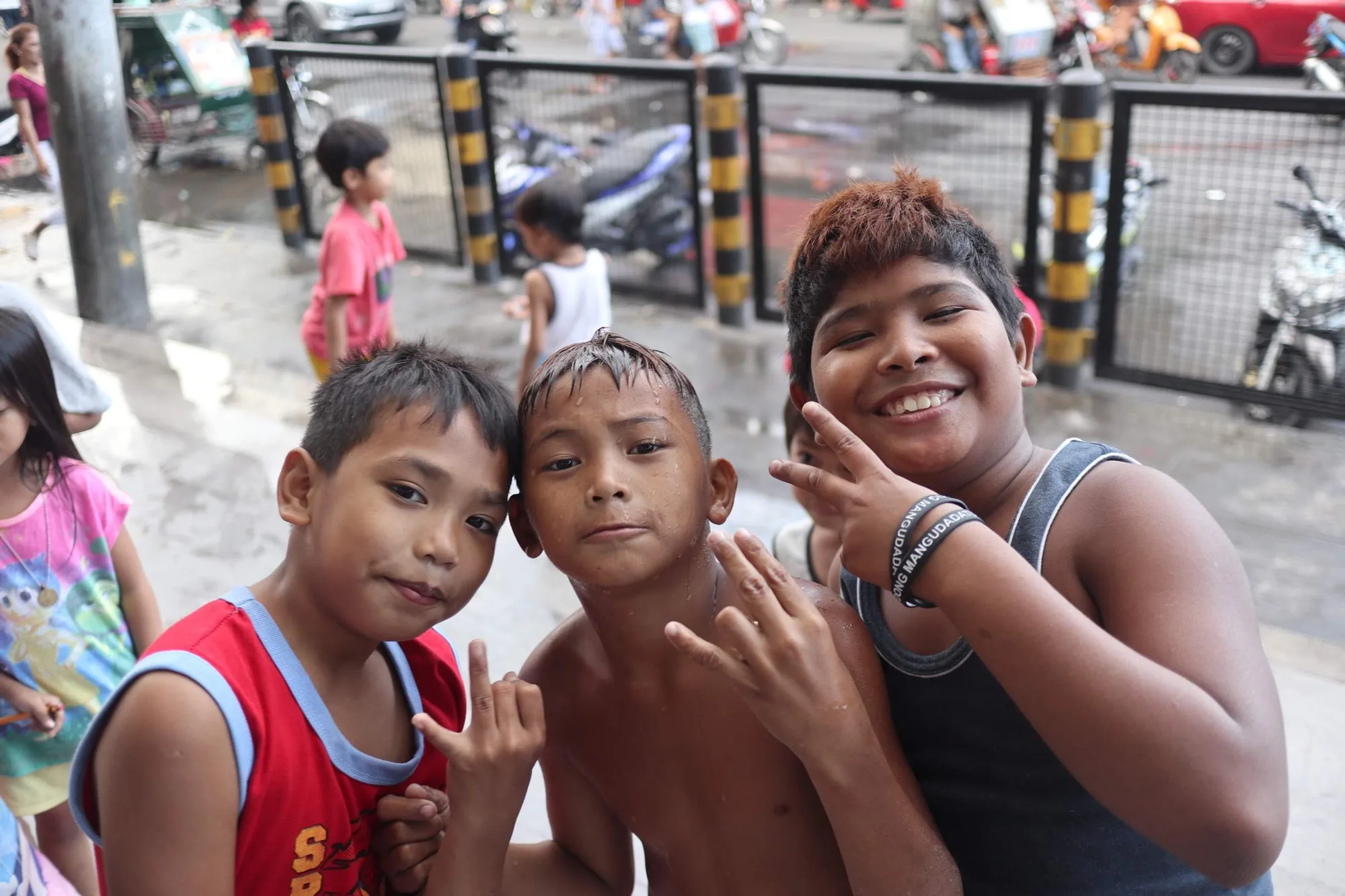 フィリピンのマニラで撮影された写真です。写真には、手前に3人の少年が写っており、皆カメラに向かって笑顔を見せています。一番右の少年は笑顔でピースサインをしています。他の少年もそれぞれ異なるポーズをとっています。3人の少年は肌の色が浅黒く、背景にはフェンス、道路、バイク、車、そして他の人々が写っています。少年たちは、ストリートで元気に遊んでいるようです。 写真全体からは、子どもたちの活気と街の喧騒が伝わってきます。