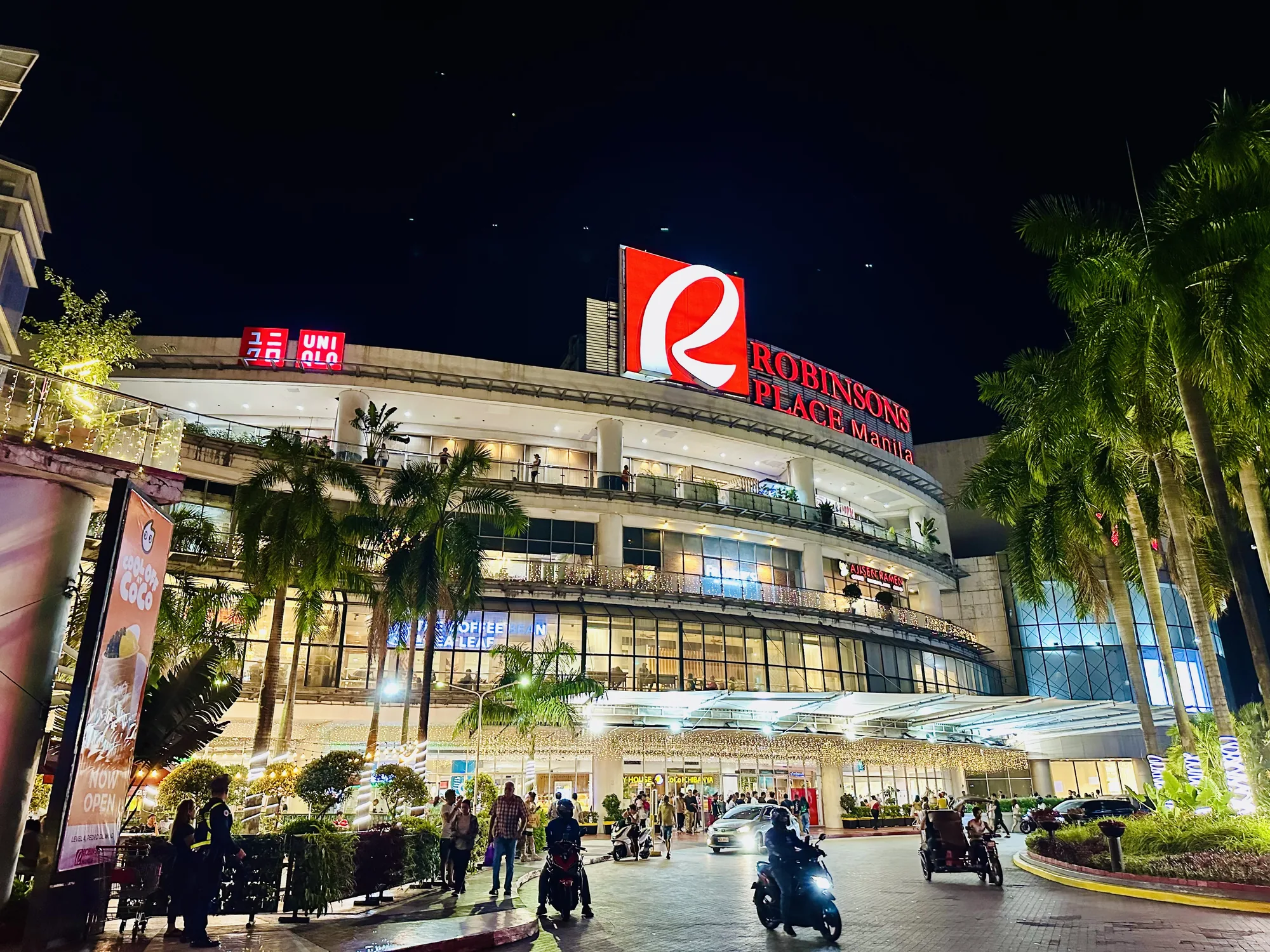 フィリピンのマニラで撮影された夜景の写真です。画面中央には、ライトアップされた大きなショッピングモール「Robinsons Place Manila」が写っています。建物の屋上には、赤い大きな「R」のロゴと「ROBINSONS PLACE Manila」の文字が見えます。建物には「UNIQLO」の看板もあります。建物の手前にはヤシの木が立ち並び、南国らしい雰囲気を醸し出しています。モール前には、歩行者やバイク、トライシクルが行き交い、賑わっています。イルミネーションが施され、全体的に華やかで活気のある印象です。広告の看板も見えます。