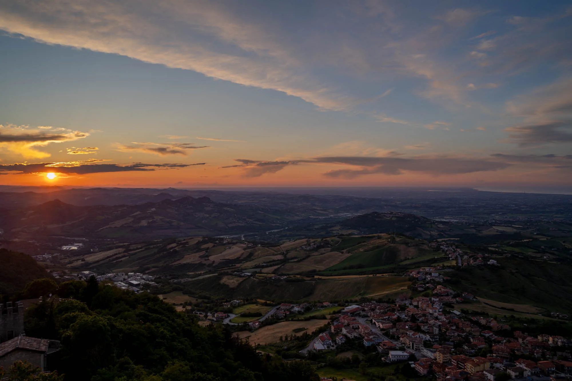 写真はサンマリノで撮影された夕暮れ時の風景です。夕日が地平線に沈みかけ、空はオレンジ色から青色へとグラデーションを描いています。雲が夕日に照らされ、金色に輝いています。眼下には緑豊かな丘陵地帯が広がり、遠くまで見渡せる素晴らしい眺めです。手前には赤い屋根の家々が密集した村が見え、夕日に照らされて温かい光を放っています。丘陵地帯には畑や道路が走り、人の営みが感じられます。全体的に、夕日の美しさと自然の雄大さが調和した、静かで美しい写真です。