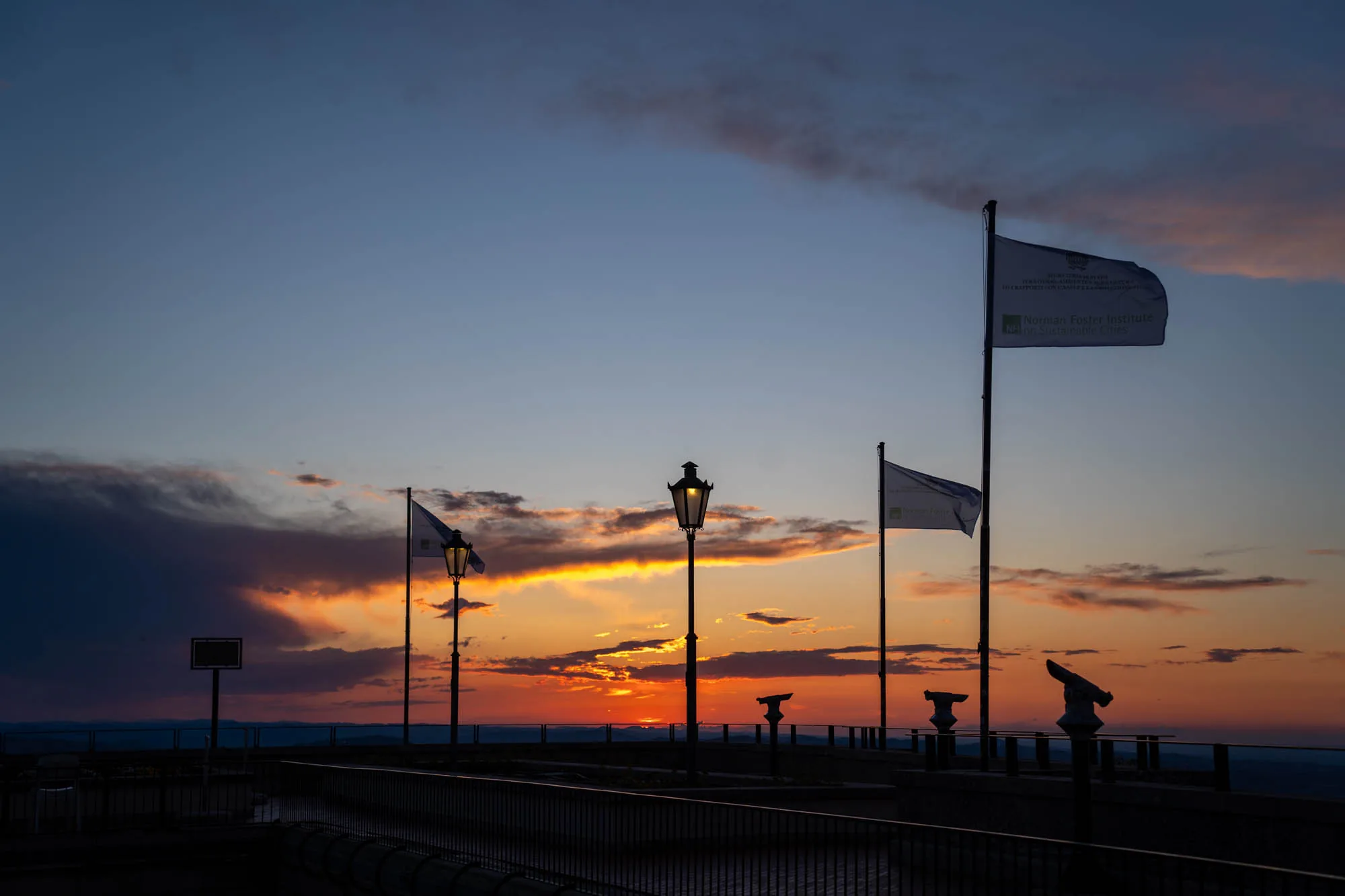 写真は、サンマリノで撮影された夕焼けの風景を捉えています。手前には、柵で囲まれた展望台のような場所があり、そこには古いスタイルの街灯が立っています。街灯はシルエットになっており、その光がわずかに灯っています。さらに、双眼鏡型の望遠鏡がいくつか設置されています。画面奥には、夕焼け空が広がり、オレンジ色やピンク色の光が雲を染めています。空の一部は濃いグレーの雲で覆われており、ドラマチックな雰囲気を醸し出しています。写真には、旗が3本立っており、そのうちの1本には「Norman Foster Institute」という文字が見えます。全体的に、静かで美しい、そして少し神秘的な印象を与える写真です。