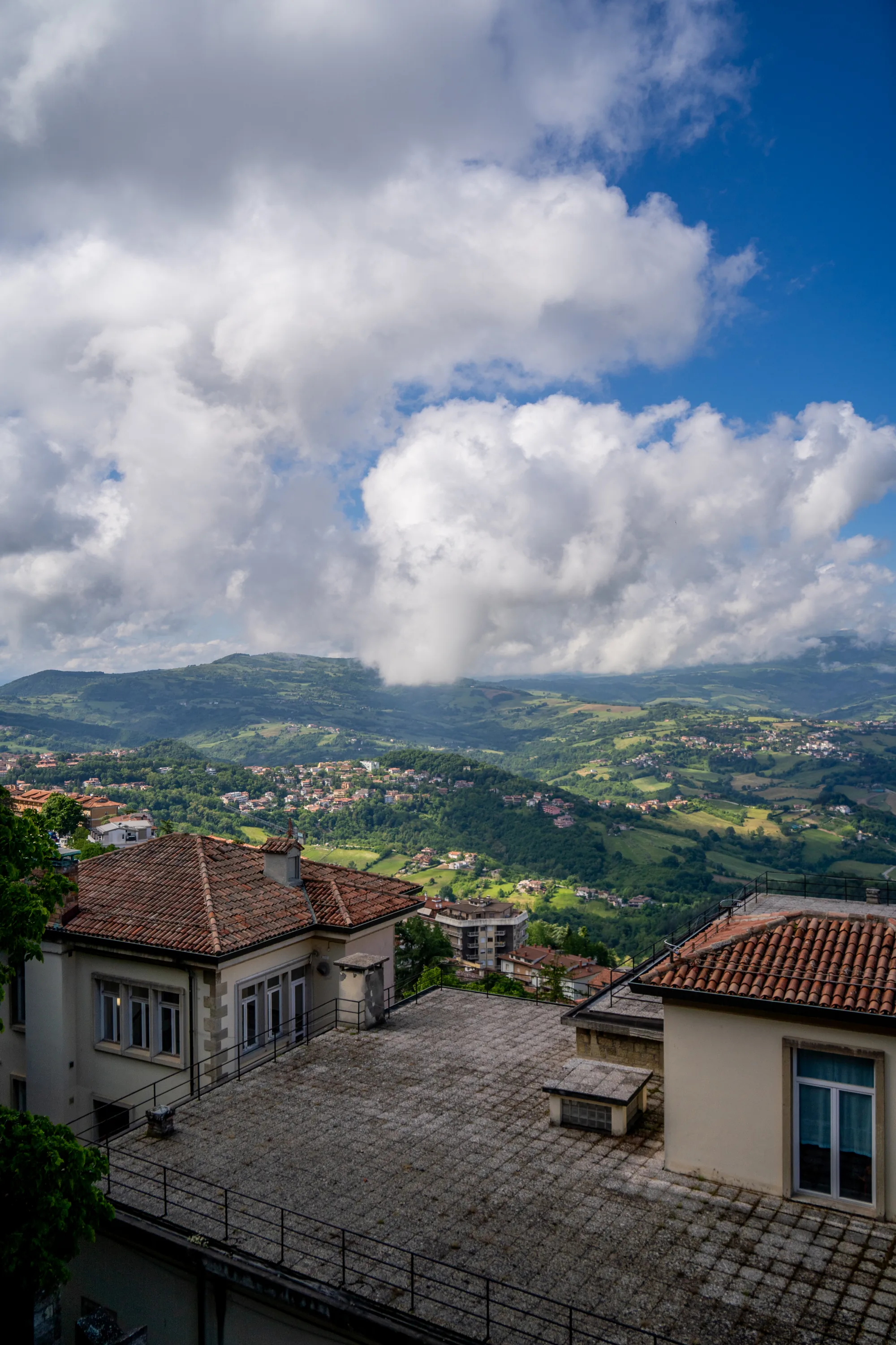 写真はサンマリノで撮影されたもので、建物の上から遠くの風景を見渡す構図となっています。手前には、オレンジ色の瓦屋根の建物が見え、その屋上は灰色がかったタイルで覆われています。屋上には黒い柵が設けられています。  背景には、緑豊かな丘陵地帯が広がっており、遠くには集落や町並みが点在しています。空には、大きな白い雲が浮かび、青空とのコントラストが美しいです。雲は山々に影を落としており、奥行きを感じさせます。全体的に、明るく開放的な雰囲気の写真です。風景写真として、サンマリノの美しい自然と建物の調和が捉えられています。