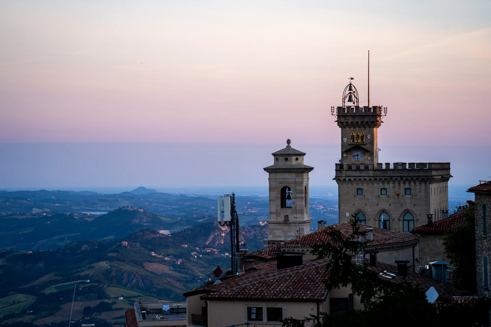写真はサンマリノで撮影されたもので、夕暮れ時の風景を捉えています。空はピンクと紫のグラデーションに染まり、遠くまで見渡せる山々の景色が広がっています。手前には、赤茶色の屋根が連なる建物群があり、その上に鐘楼や時計塔がそびえ立っています。時計塔には、時計の文字盤と3人の人物像が飾られています。また、通信アンテナも立っており、歴史的な建造物と現代的な設備が共存している様子がわかります。全体的に落ち着いた雰囲気で、夕暮れの静けさとサンマリノの美しい景観が感じられる写真です。