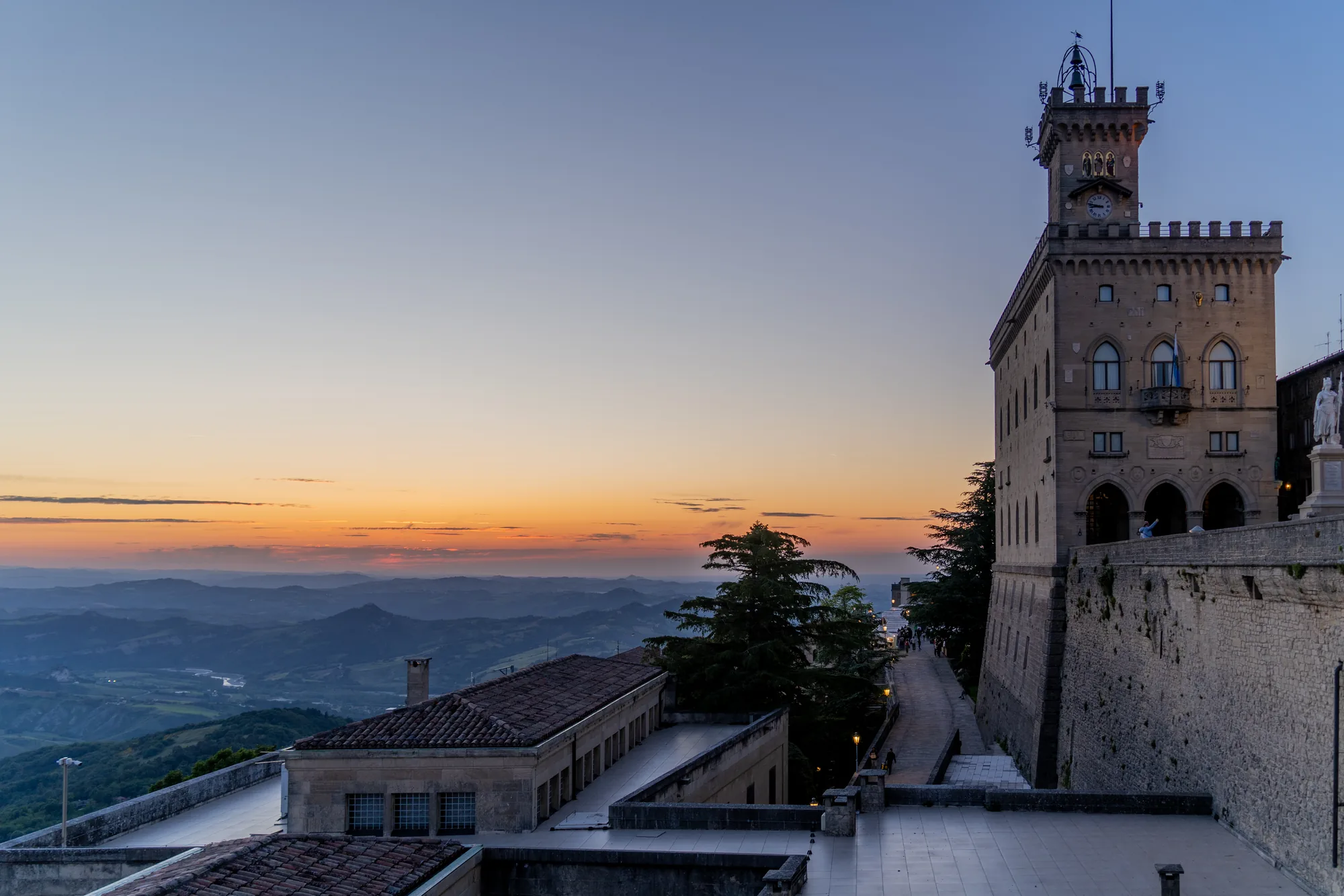 写真はサンマリノで撮影された夕景である。画面奥にはオレンジ色に染まる空と、遠くの山々が連なっている。手前には中世の建物がそびえ立っており、特に右側の高い塔が印象的だ。塔には時計が設置され、建物の壁は石造りで歴史を感じさせる。建物の間には通路があり、観光客らしき人々の姿が見える。低い建物が手前にあり、オレンジ色の空とのコントラストが美しい。写真全体としては、サンマリノの歴史的な建造物と夕暮れの美しい風景が組み合わさった、静かで落ち着いた雰囲気である。