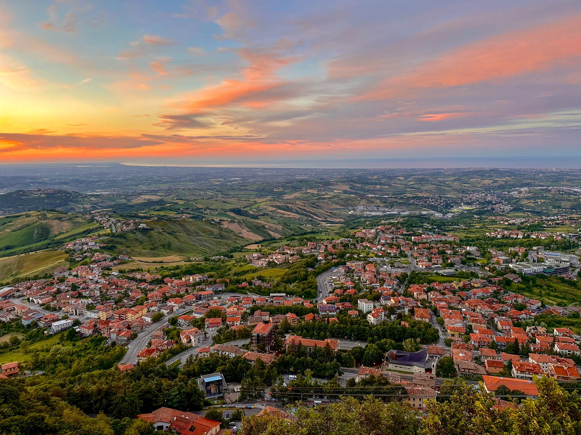 写真は、サンマリノ共和国と思われる場所から撮影された風景を捉えています。空は、オレンジ色からピンク、そして薄い青へとグラデーションを描き、夕暮れ時の美しい光景を作り出しています。雲は薄く広がり、空の色彩をさらに豊かにしています。  遠くには地平線が見え、おそらく海が広がっています。その手前には、緑豊かな丘陵地帯が広がっており、畑や森、そして点在する集落が確認できます。  手前の地域には、赤茶色の屋根を持つ建物が密集しており、それが集落を形成していることがわかります。道路が建物と建物の間を縫うように走っており、その様子から、この場所が生活の場であることが伝わってきます。写真全体を通して、自然の美しさと人々の営みが調和している様子が感じられます。