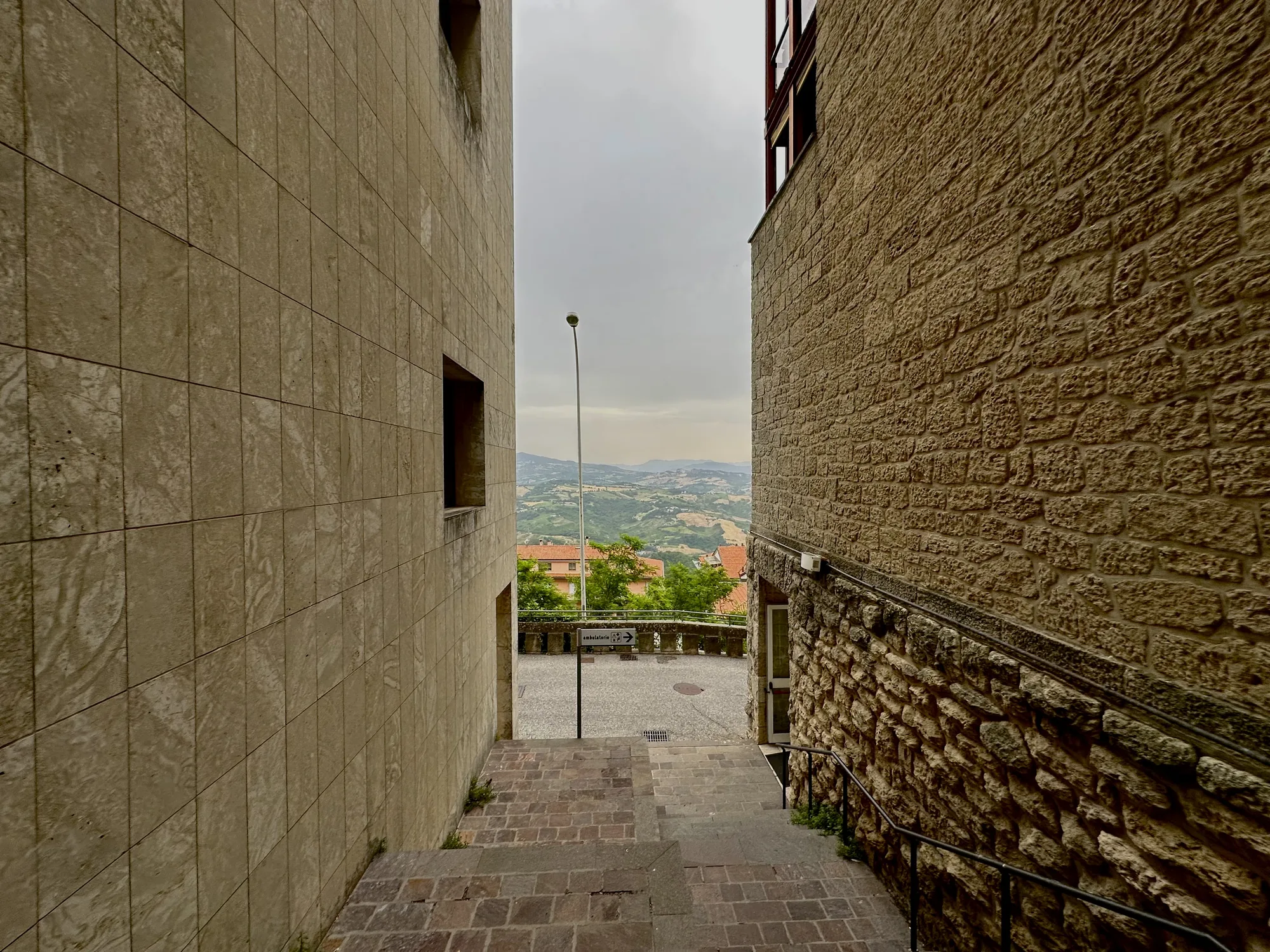 サンマリノで撮影された写真です。石畳の階段が左右の建物に挟まれた細い道を進んでいます。左側の建物は、綺麗に並んだ石材で覆われており、窓がいくつか見えます。右側の建物は、粗い石積みの壁で、歴史を感じさせます。階段を下った先には広場があり、その奥には緑豊かな丘陵地帯が広がっています。空は曇り空で、全体的に落ち着いた雰囲気です。道案内を示す看板も見え、観光地の風景であることが伺えます。建物の石の色合いと風景の緑が調和した、美しい一枚です。