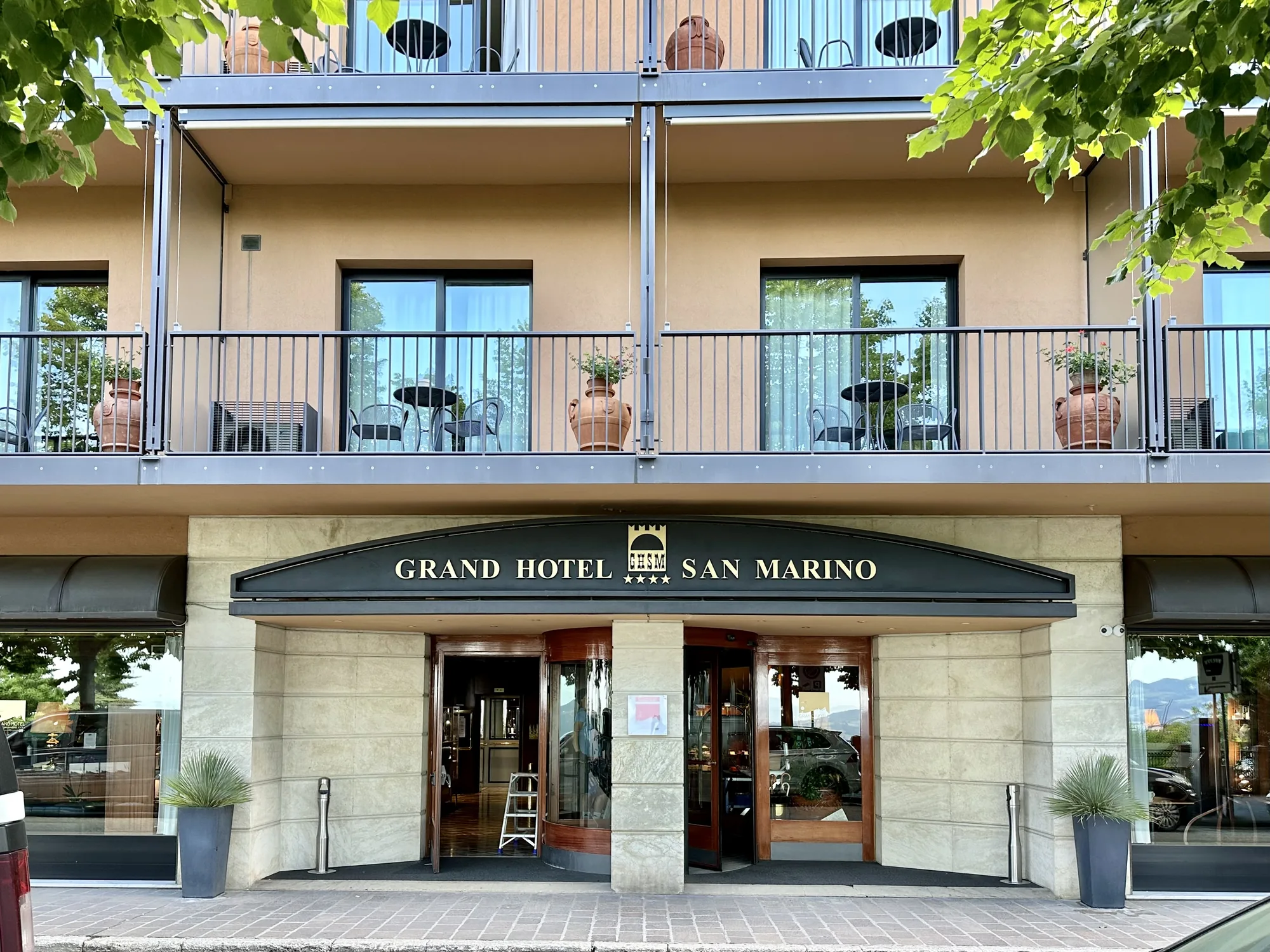 写真には、サンマリノにある「GRAND HOTEL SAN MARINO」の外観が写っています。建物はベージュの色調で、2階建て構造に見えます。各階にはバルコニーがあり、テーブルと椅子が配置され、テラコッタのプランターが飾られています。建物の入り口には、「GRAND HOTEL SAN MARINO」と書かれた看板があり、星のマークが４つ付いています。入り口は回転ドアと通常のドアで構成され、どちらも開いています。左側のドアの奥には、梯子が見えます。入り口の両側には、スタイリッシュな植木鉢に入った植物が飾られています。写真の右側には緑の木々が部分的に写り込んでおり、建物の奥には丘陵地帯が遠くに見えます。全体的に、上品で落ち着いた雰囲気の写真です。