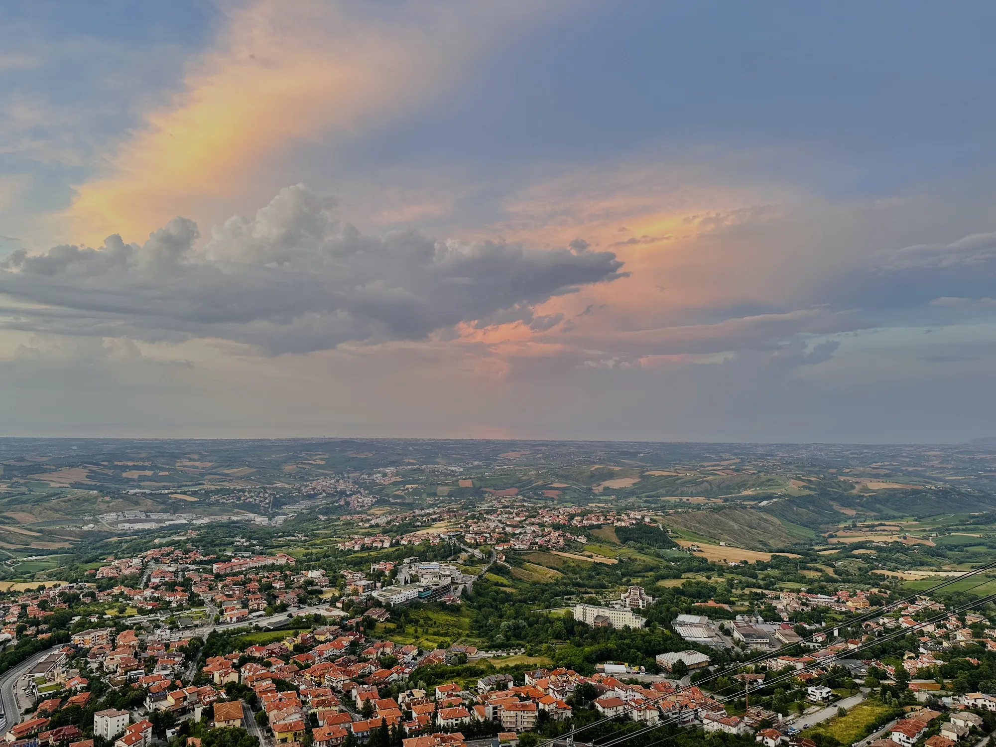 この写真はサンマリノで撮影された風景写真です。高い場所から見下ろした景色が広がっており、遠くまで見渡せる構図になっています。空には、灰色やピンクが混ざった雲が広がり、夕焼けのような淡い光が差し込んでいます。地平線付近は霞んでおり、幻想的な雰囲気を醸し出しています。  手前には、オレンジ色の屋根が特徴的な家々が密集した街並みが広がっています。家々の間には緑豊かな木々が生い茂り、自然と都市が調和した景観を作り出しています。さらに遠方には、丘陵地帯が広がり、畑や森林がパッチワークのように配置されています。遠くには海らしきものも見えます。全体的に落ち着いた色調で、穏やかで美しいサンマリノの風景を捉えた写真です。