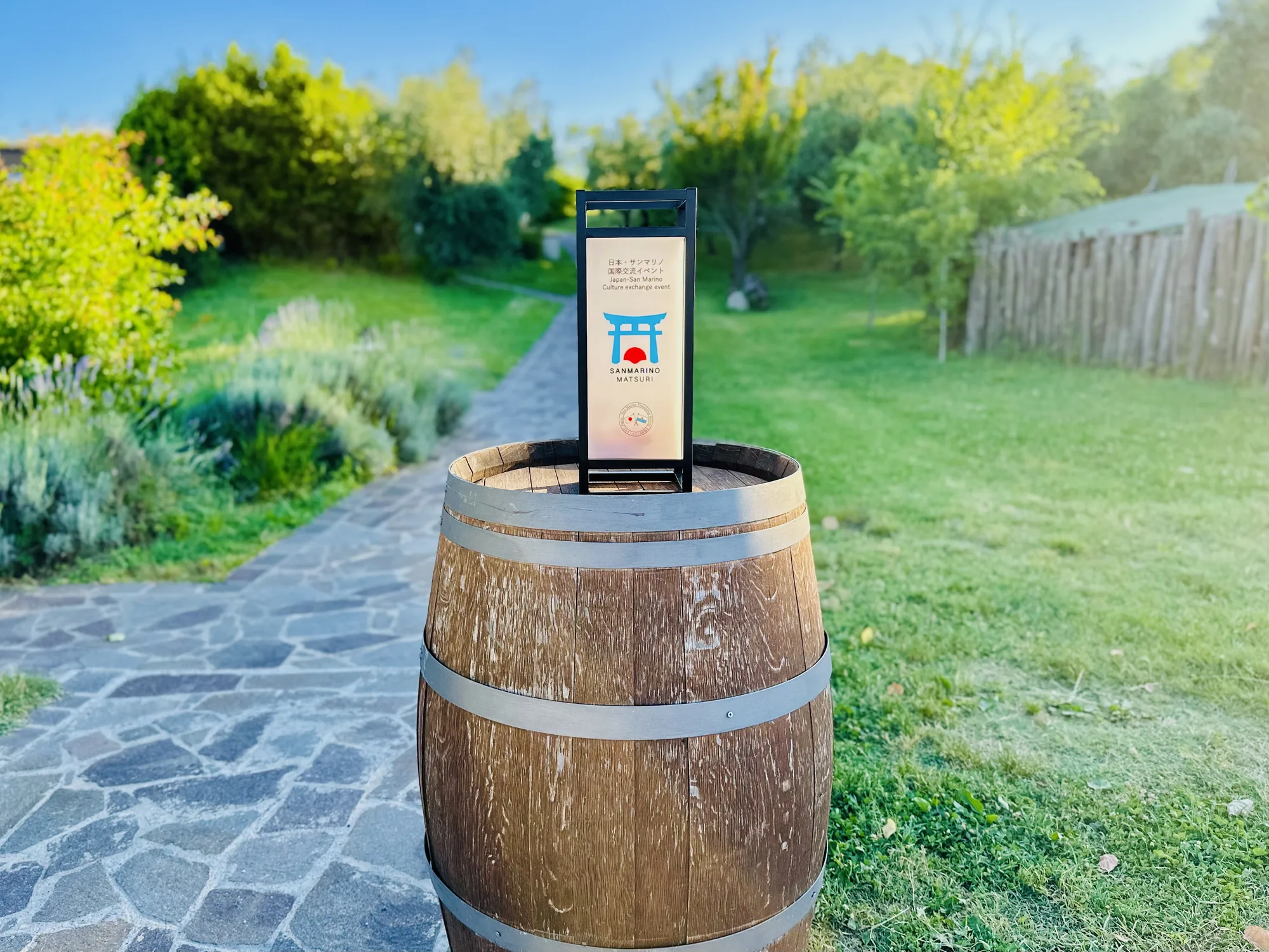 写真には、木製の樽の上に立てられた、サンマリノ祭りのイベント告知看板が写っています。看板には「日本・サンマリノ国際交流イベント」と日本語と英語で書かれており、鳥居と日の丸を組み合わせたようなロゴマークが描かれています。背景には緑豊かな庭園が広がり、石畳の小道が奥へと続いています。庭園には木々や草花が生い茂り、穏やかな雰囲気です。全体的に明るく、平和な印象を与える写真です。