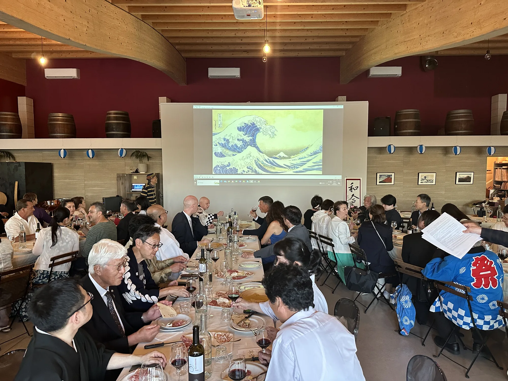 写真は、長テーブルを囲んで多くの人が食事をしている様子を捉えています。壁には、日本の浮世絵である葛飾北斎の「神奈川沖浪裏」が大きく投影されています。参加者はワイングラスを手に持ち、テーブルにはワインボトル、ハムやパンなどの軽食が並んでいます。会場は木製の梁がむき出しになった天井や、壁に飾られた酒樽など、独特の雰囲気を持っています。一部の参加者は和装をしており、法被を着用している人もいます。室内の装飾には提灯も使われており、全体的に和のテイストが感じられる食事会であることがわかります。