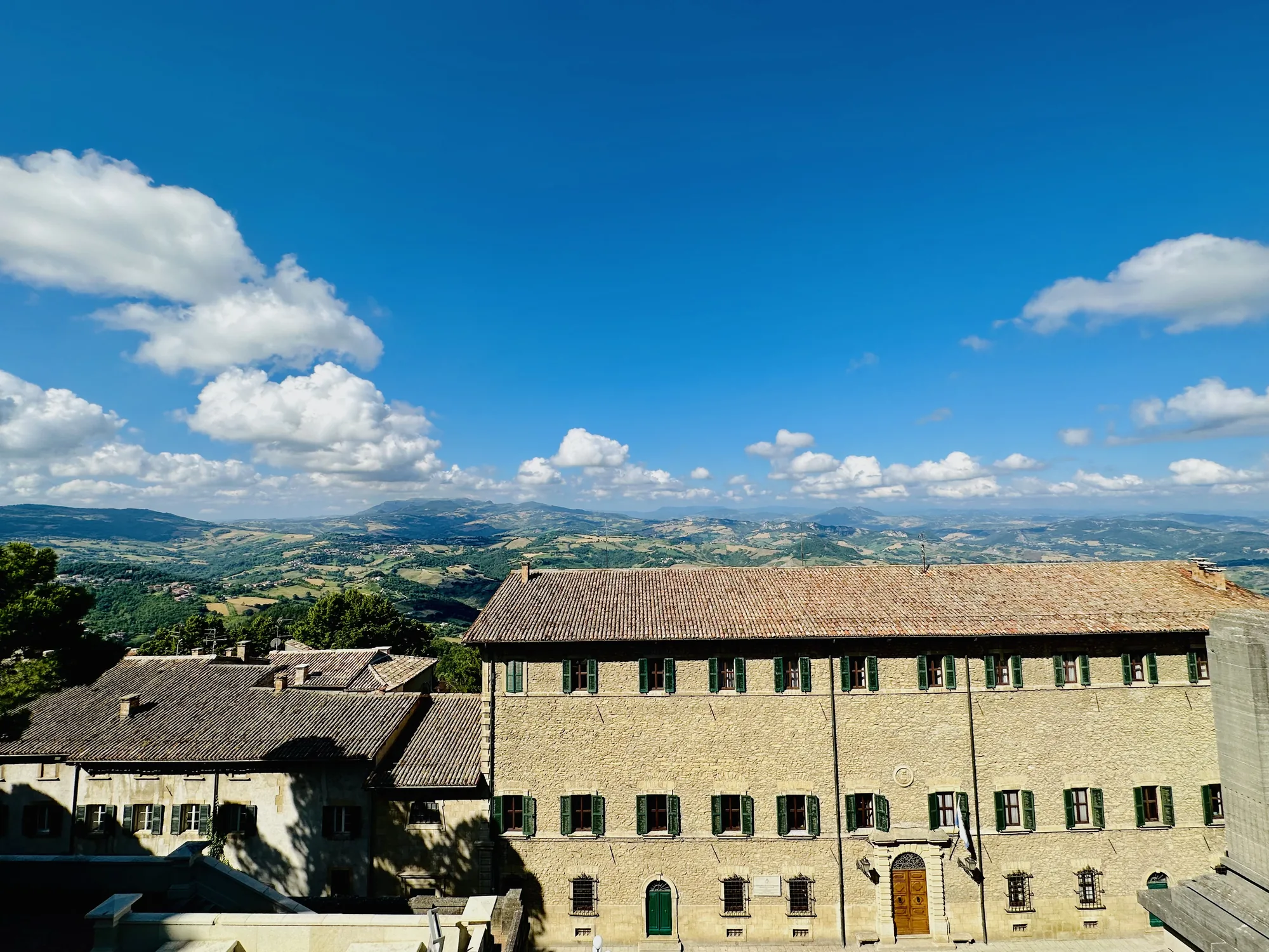 写真は、サンマリノの建物と周囲の風景を捉えたものです。空は青く、白い雲が点在しており、遠くに緑豊かな丘陵地帯が広がっています。手前には、石造りの建物が写っており、茶色の屋根瓦と緑色の窓枠が特徴的です。建物の壁は、太陽の光を浴びて明るく照らされています。建物の手前には、別の建物の一部が見え、その影が落ちています。全体的に、穏やかで美しい風景が広がっており、サンマリノの歴史的な雰囲気と自然の豊かさを感じさせる写真です。