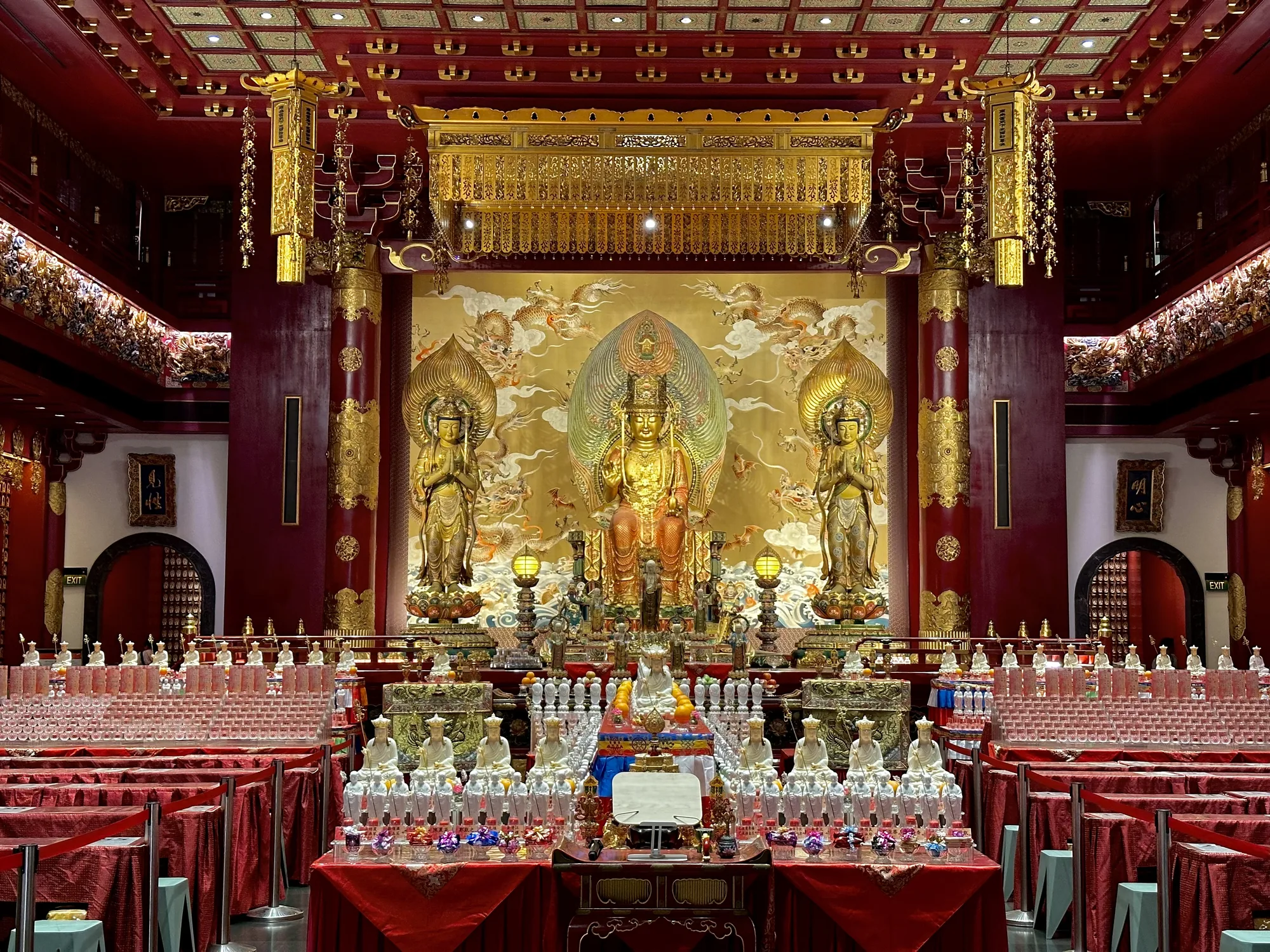 写真は、シンガポールの寺院内部を撮影したものです。全体的に赤と金の配色が印象的で、豪華絢爛な雰囲気を醸し出しています。正面には、金色の大きな仏像が安置されており、その背景には龍の絵が描かれています。仏像の両脇には、それぞれ2体の像が立っています。天井は格子状になっており、金色の装飾が施されています。手前には、赤い布がかけられたテーブルが整然と並んでおり、その上には蝋燭立てや小さな仏像、そして多数の水の入ったグラスが置かれています。壁には、出口を示す「EXIT」の文字が見えます。全体として、寺院の荘厳さと信仰心が感じられる一枚です。