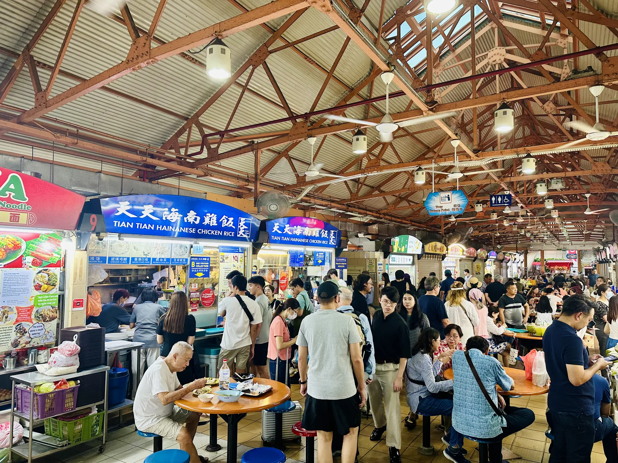 写真はシンガポールの活気ある屋台街の様子を捉えています。天井は古い鉄骨構造で、そこから多数の扇風機と照明が吊り下げられています。画面奥には、多くの人々が食事をしたり、列に並んだりしている様子が伺えます。手前には、「Tian Tian Hainanese Chicken Rice」と書かれた青い看板の屋台が見えます。その他にも様々な屋台が並び、多様な料理を提供しているようです。人々はカジュアルな服装をしており、テーブルを囲んで談笑したり、一人で食事を楽しんだりしています。全体的に賑やかで、ローカルな雰囲気が感じられる写真です。