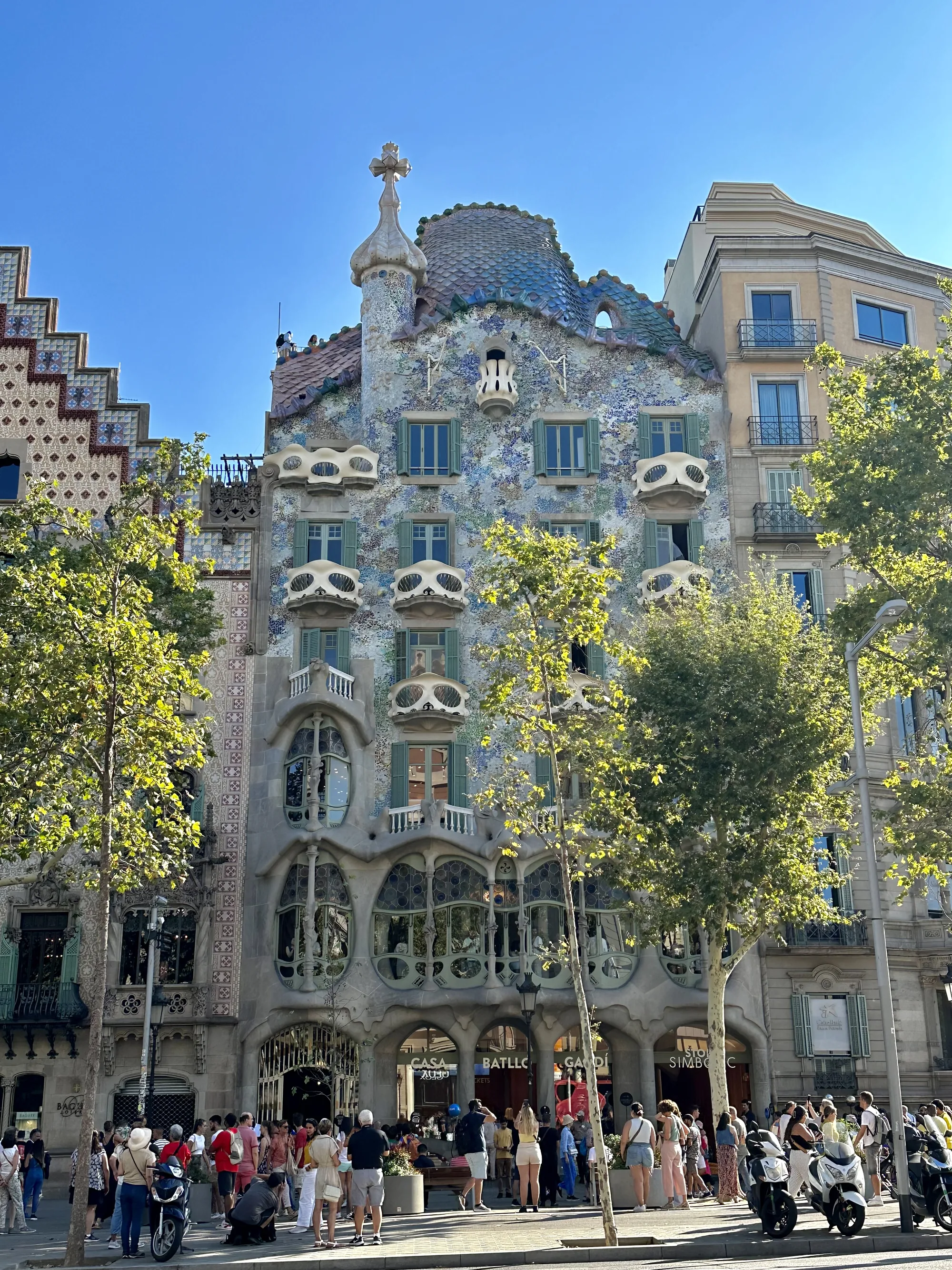 写真はスペインのバルセロナで撮影されたもので、アントニ・ガウディの代表作であるカサ・バトリョの外観を捉えています。青空を背景に、特徴的な波打つようなファサードが印象的です。建物全体はモザイクタイルで覆われており、青や緑、白などの色が組み合わさって独特の雰囲気を醸し出しています。窓は様々な形をしており、骸骨の口のようなバルコニーが目を引きます。屋根は鱗のようなデザインで、頂上には十字架が立っています。建物の前には観光客が集まっており、建物の壮大さを目の当たりにしている様子がうかがえます。建物の左右には街路樹が植えられており、日差しを遮っています。写真全体を通して、ガウディ建築の独創性とバルセロナの活気が感じられます。