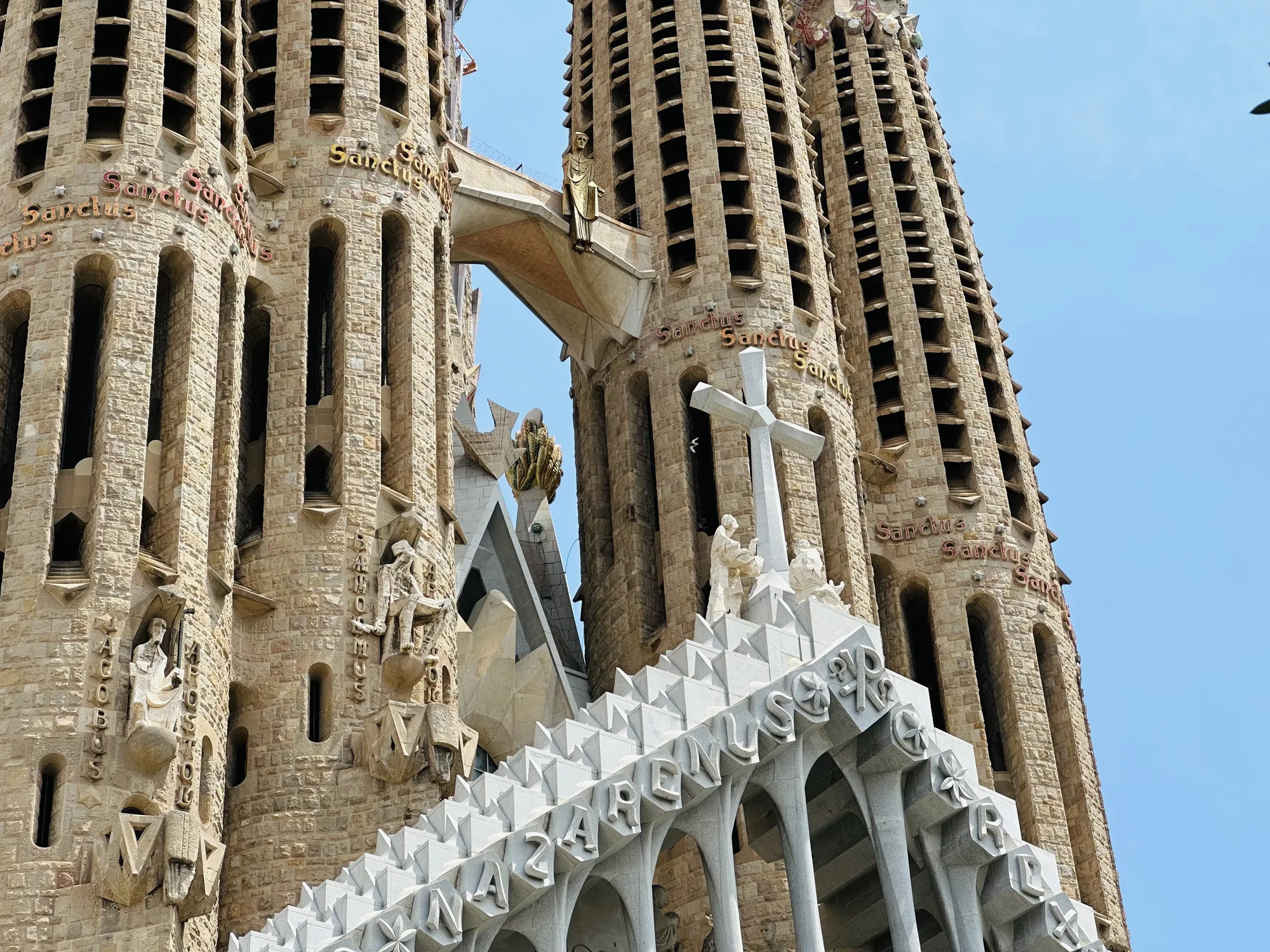 写真は、スペインのバルセロナにあるサグラダファミリア教会の細部を捉えたものです。カメラは下から上を見上げるように構図されており、建物の印象的な高さと複雑なデザインを強調しています。教会の外観は、長い年月を経た石造りで、窓や装飾が施された塔がそびえ立っています。塔には「Sanctus」の文字が刻まれています。 また、白い石で作られた、幾何学的な形状とアーチを持つ部分も写っており、頂上には十字架と人物像が配置されています。空は明るい青色で、教会の石の色と美しいコントラストをなしています。全体的に、写真からは壮大さ、宗教性、そしてガウディ建築の独創性が伝わってきます。