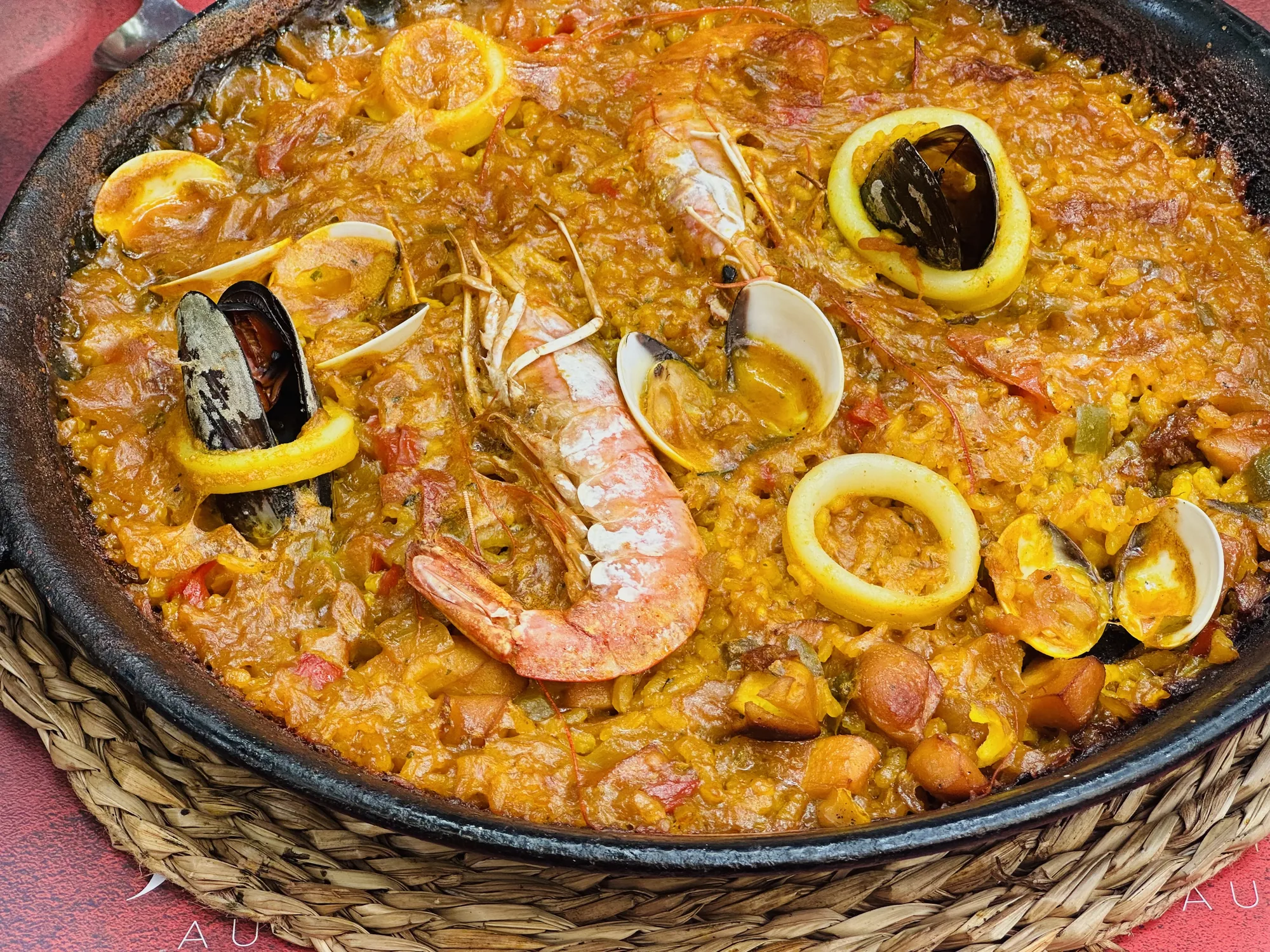 スペインのバルセロナで撮影された、食欲をそそるパエリアの写真です。大きな黒いパエリア鍋には、サフランライスをベースに、エビ、ムール貝、アサリ、イカなどの新鮮なシーフードがたっぷりと盛り付けられています。ご飯はオレンジ色に輝き、魚介の旨味が凝縮されていることが伝わってきます。イカは輪切りにされ、ムール貝やアサリは殻付きで提供されており、見た目にも華やかです。鍋は藁のような素材で編まれたコースターの上に置かれ、温かみのある雰囲気を醸し出しています。全体的に、本格的なスペイン料理の美味しさを表現した、魅力的な一枚です。