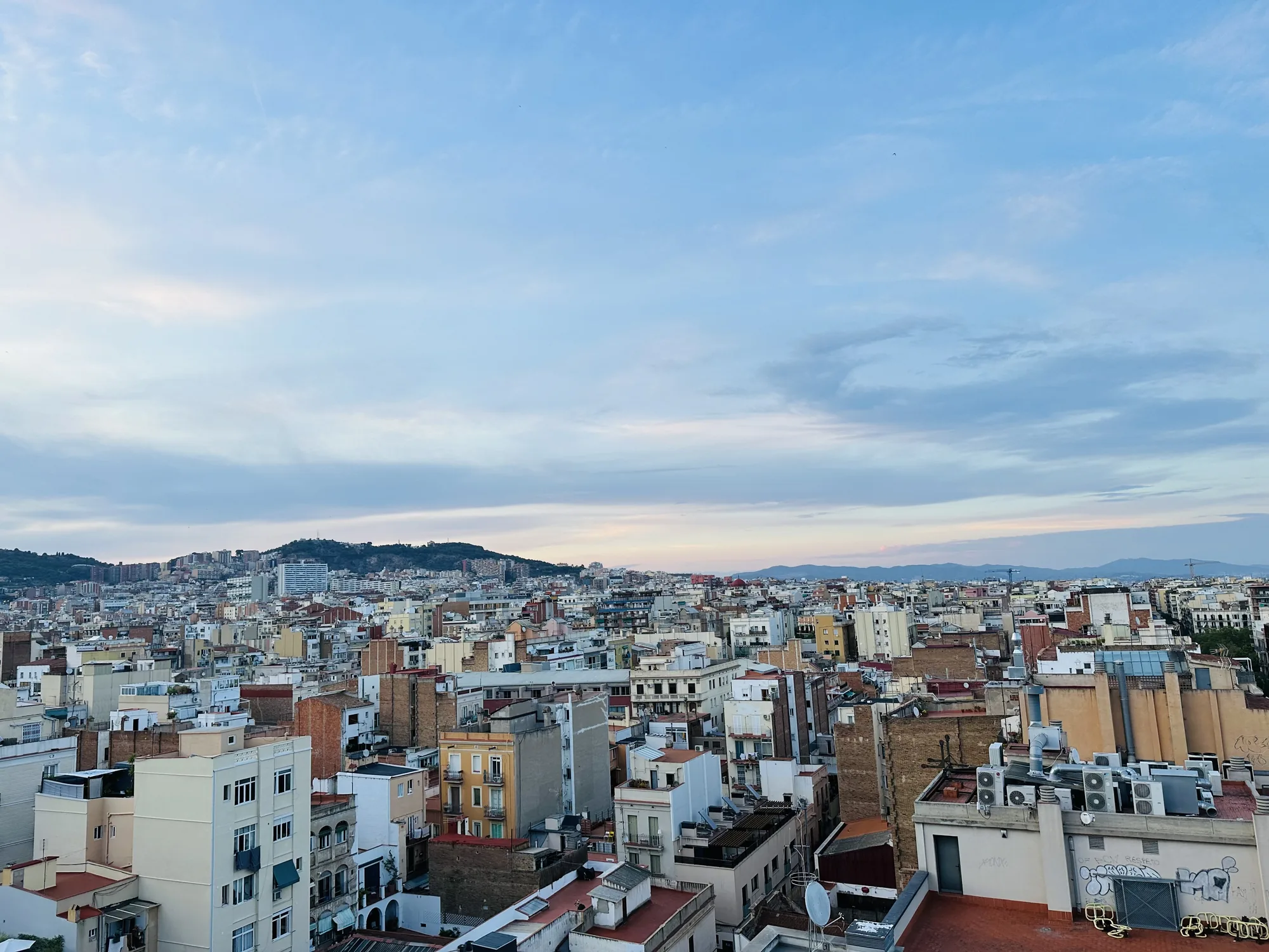写真はスペイン、バルセロナの街並みを捉えたものです。高台から見下ろすように撮影されており、密集した建物群が一面に広がっています。建物の多くは低層で、ベージュや白、レンガ色など落ち着いた色合いで統一されています。屋上にはエアコンの室外機やアンテナが並び、生活感を感じさせます。遠景には緑の山が連なり、街の風景に自然の要素を加えています。空は淡い青色で、薄い雲が広がっており、穏やかな天候であることが伺えます。全体的に静かで落ち着いた雰囲気の写真です。