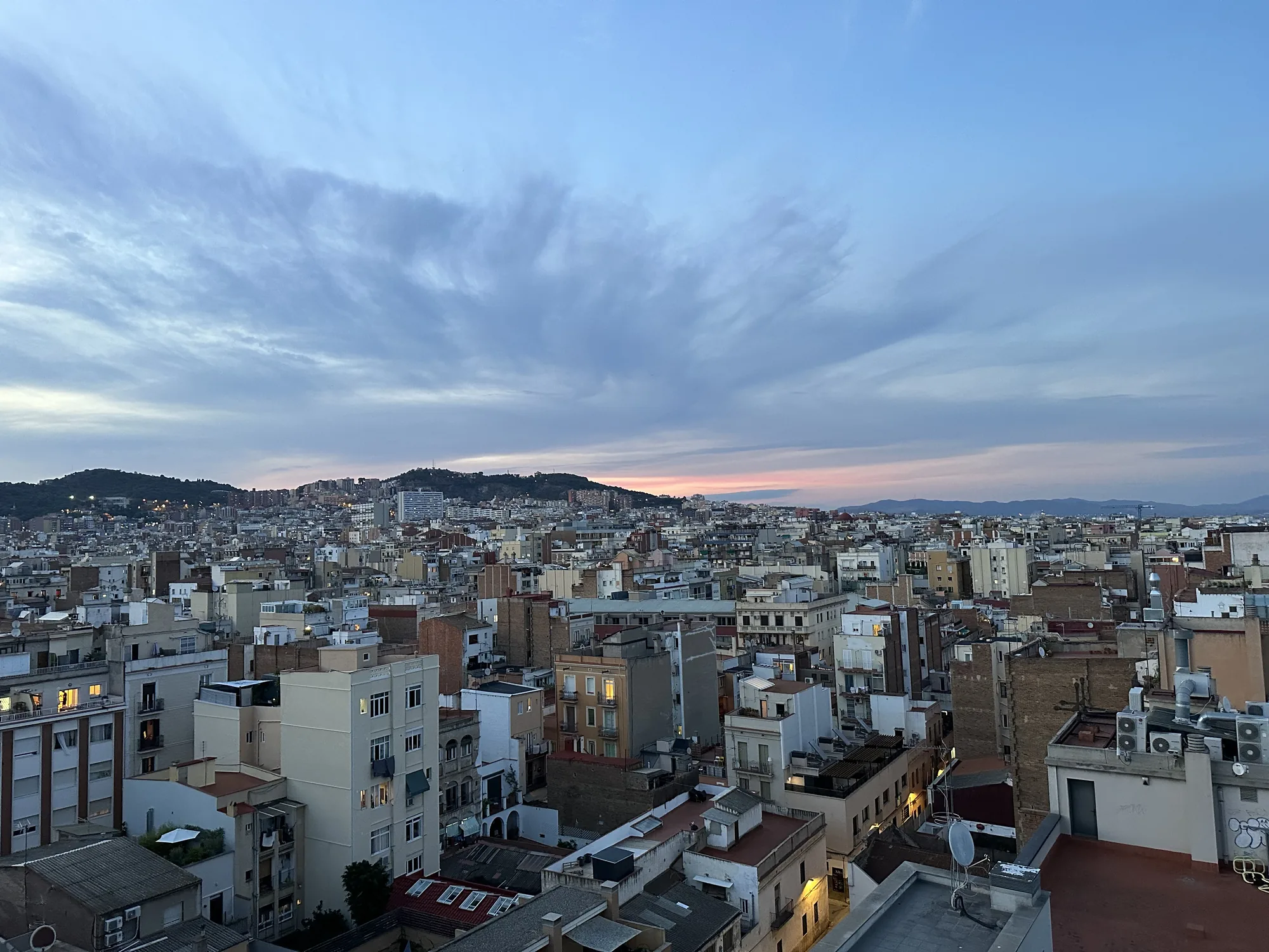 写真はスペイン、バルセロナの市街地を上空から捉えたものです。夕暮れ時の空は淡いピンク色から青色へとグラデーションを描き、雲が広がり、街並みを優しく照らしています。眼下には密集した建物が広がり、伝統的な赤茶色の屋根瓦と白い壁が特徴的な建物が目立ちます。いくつかの窓には明かりが灯り始めており、生活の温かさを感じさせます。遠くには山並みが見え、市街地の背景として奥行きを加えています。全体的に穏やかで静かな雰囲気の写真です。