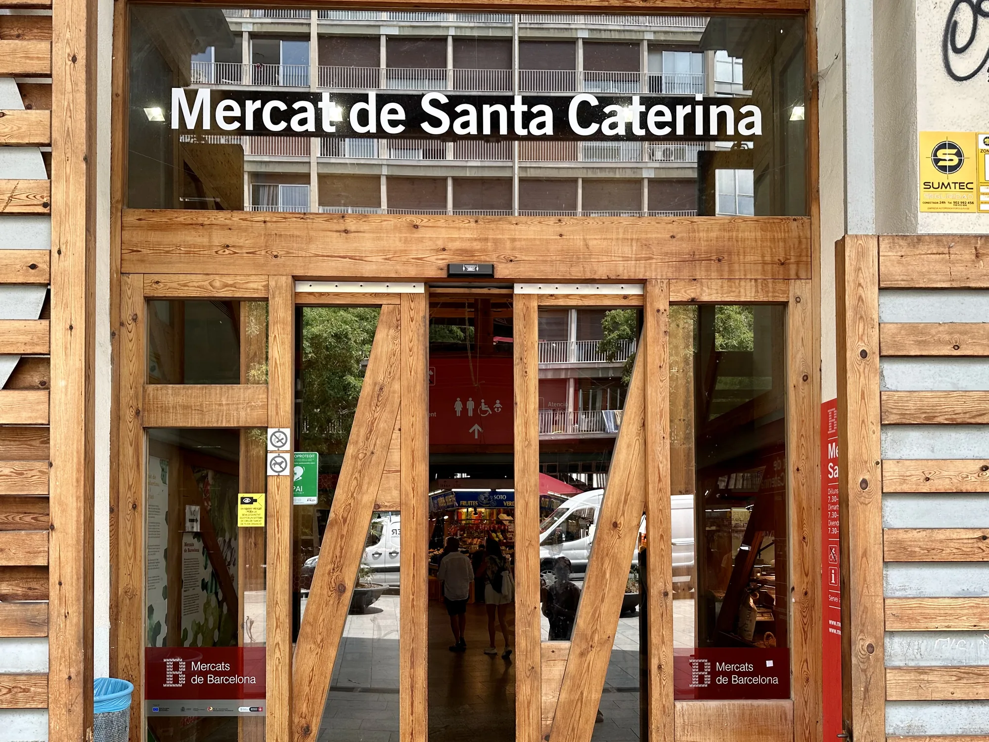この写真は、スペインのバルセロナにあるサンタ・カテリーナ市場の入口を捉えたものです。木製のユニークなデザインのドアが特徴的で、ドアの上部には「Mercat de Santa Caterina」という市場の名前が書かれています。ドアのガラス越しには、市場内部の様子がぼんやりと見え、買い物客や商品の一部が写っています。市場の向かいには、集合住宅のような建物が見えます。ドアには、営業時間や市場に関する案内などが記載されたステッカーや看板が貼られています。全体的に活気があり、地元の人々や観光客に親しまれている市場の雰囲気が伝わってきます。