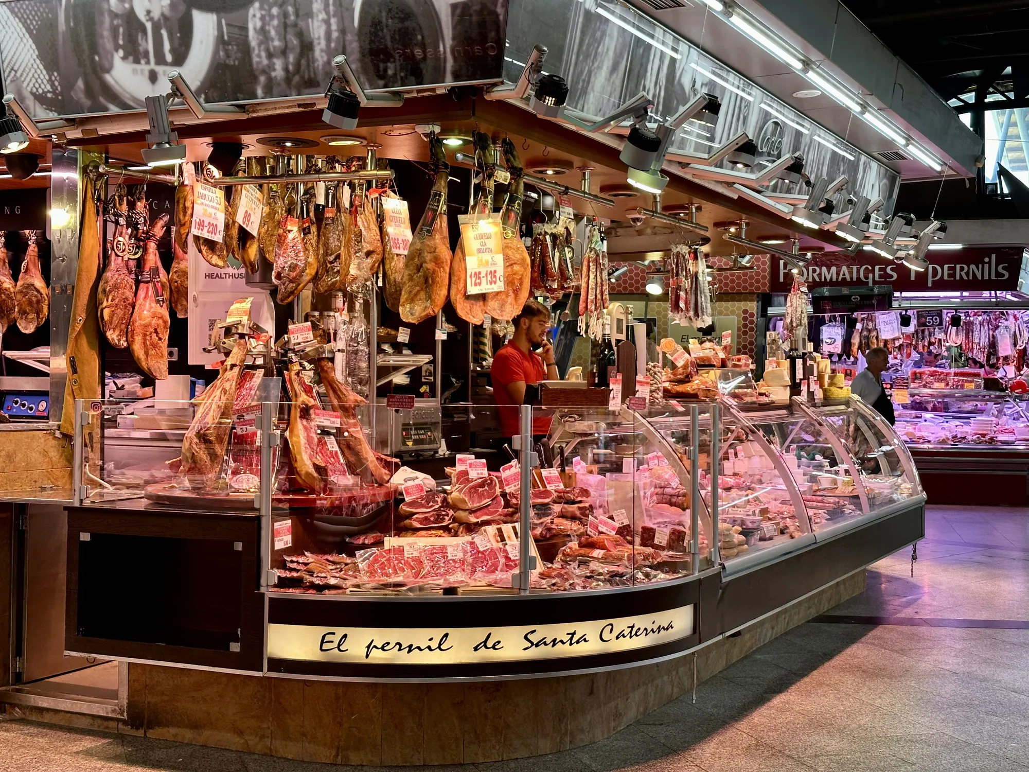 写真はスペインのバルセロナで撮影された、賑やかな市場の様子を捉えています。特に焦点が当てられているのは、豊富な種類の生ハムを販売する「El pernil de Santa Caterina」という名前の店です。天井からは数多くの生ハムの塊が吊り下げられ、それぞれに価格が書かれた札がついています。ショーケースには、スライスされた生ハムやサラミ、チーズなどが美しく陳列されています。赤色のシャツを着た店員が電話をしている様子が見えます。別の店も写り込んでおり、チーズや他の加工肉も販売されていることがわかります。市場の活気と、スペインの食文化の豊かさを感じさせる一枚です。