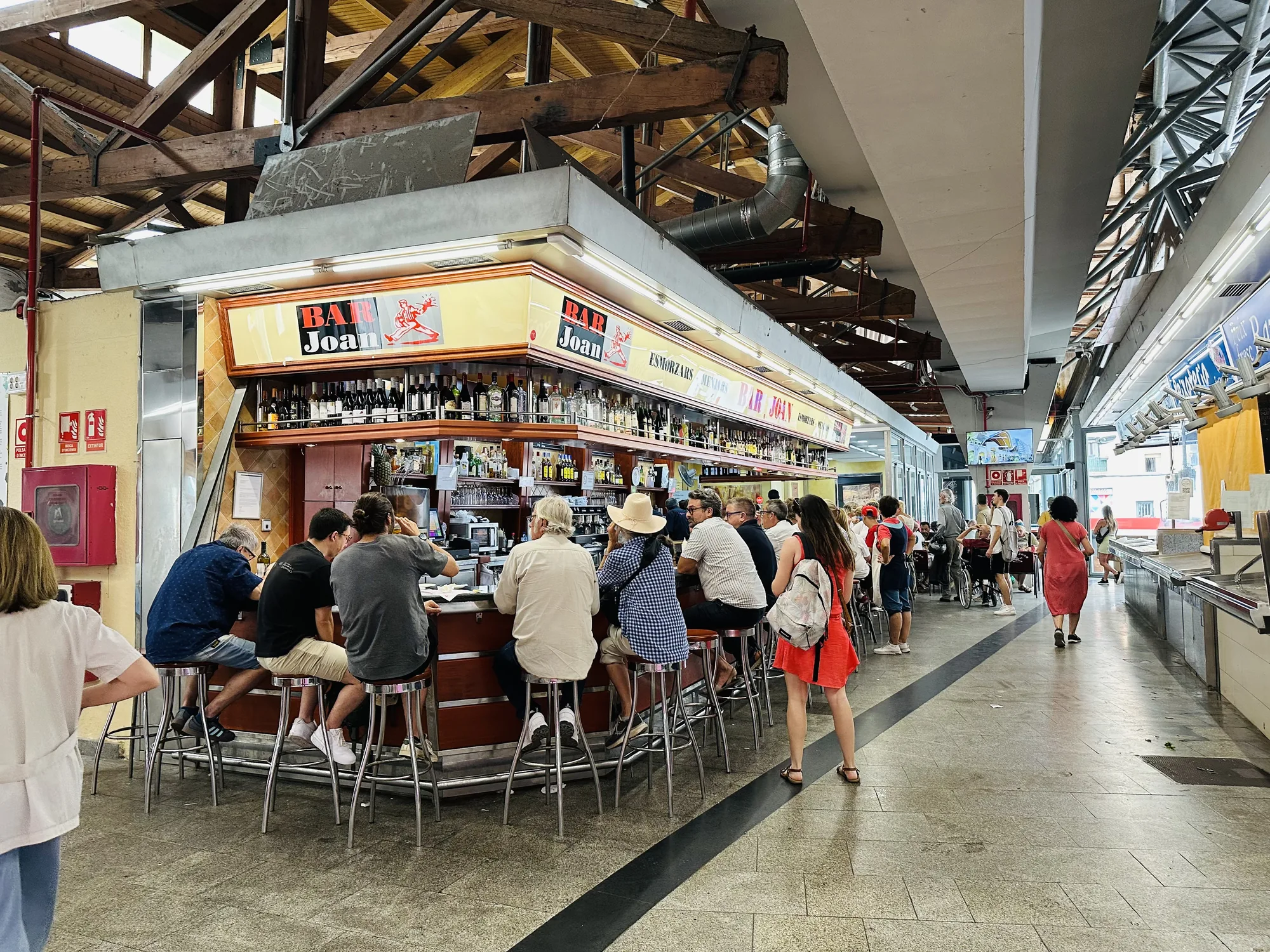 写真はスペインのバルセロナにある市場の内部を捉えたものです。中央には「BAR Joan」と書かれた賑やかなバーカウンターがあり、多くの人々がスツールに座って飲食を楽しんでいます。バーの上には、多種多様なボトルが並べられ、活気ある雰囲気を醸し出しています。天井は高く、むき出しの木製の梁が特徴的で、歴史的な建造物の一部であることを感じさせます。  通路には、買い物客や観光客が行き交い、市場の活気が伝わってきます。床は石畳で、黒い線が引かれており、通路のガイドラインとなっているようです。全体的に、市場の日常的な風景を切り取った、活気に満ちた写真です。