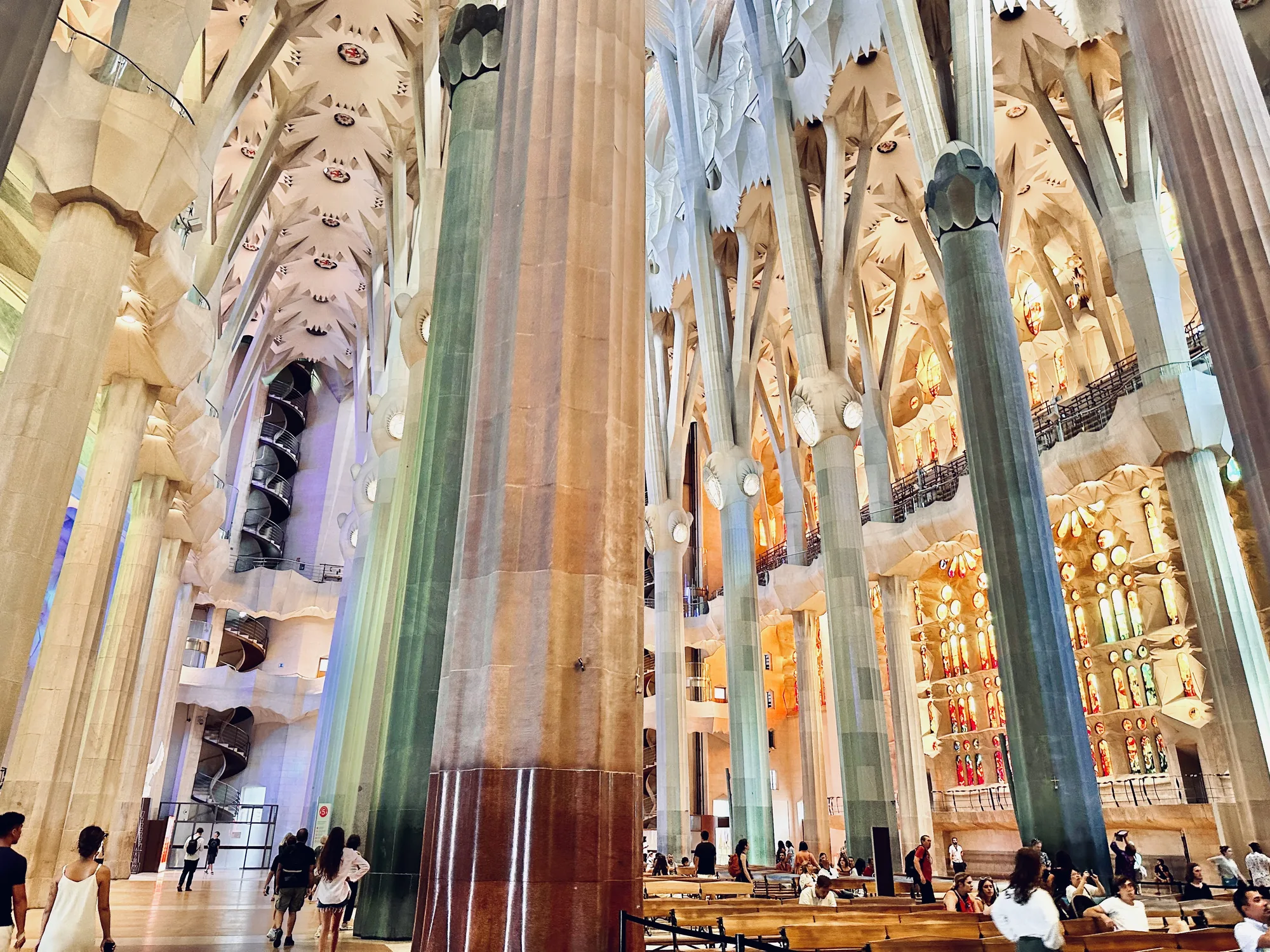 写真は、スペインのバルセロナにあるサグラダ・ファミリアの内部を捉えたものです。巨大な柱が林立し、天井に向かって伸びています。柱の色は様々で、自然光が差し込むことでさらに美しく輝いています。天井は複雑な構造で、光が散りばめられた星空のようです。ステンドグラスからは色鮮やかな光が差し込み、幻想的な雰囲気を醸し出しています。多くの観光客がベンチに座って休憩したり、写真を撮ったりして、この壮大な空間を楽しんでいる様子が伺えます。螺旋階段も見え、全体的に非常に印象的で荘厳な雰囲気です。