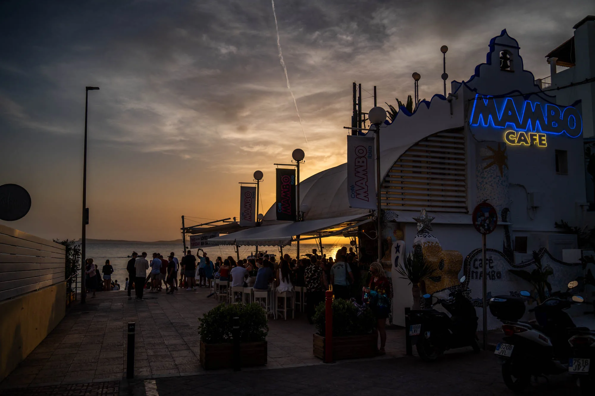 写真はスペインのイビサ島で撮影されたもので、夕暮れ時の「MAMBO CAFE」の様子を捉えています。空はオレンジ色とグレーのグラデーションで、空には飛行機雲が一本伸びています。カフェの建物は白を基調とし、青色のネオンサインで「MAMBO CAFE」と書かれています。建物の装飾は特徴的で、独特の雰囲気を醸し出しています。カフェのテラス席には多くの人が集まり、夕暮れ時のひとときを楽しんでいる様子です。店の前にはスクーターが数台停まっており、道沿いを歩く人々の姿も見えます。全体的に、夕暮れの落ち着いた雰囲気と、カフェの賑わいが感じられる写真です。